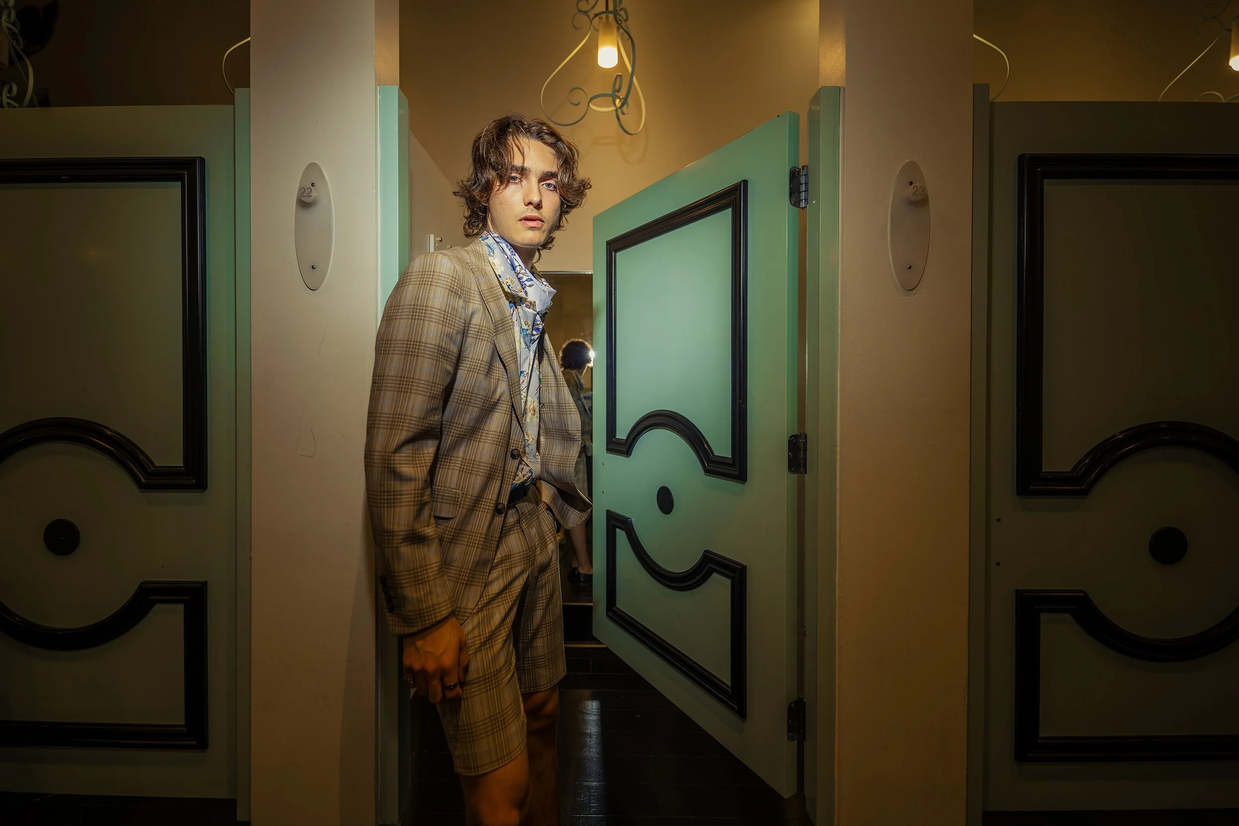 A young man with wavy hair in a plaid suit standing in front of a partially open door in a bathroom or dressing room with patterned wallpaper and warm lighting.