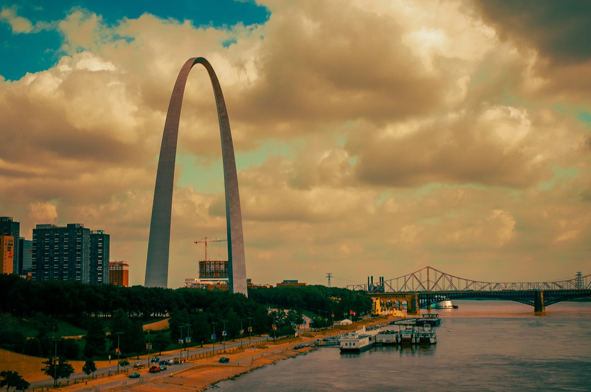 The Gateway Arch in St. Louis, Missouri, with a cloudy sky overhead, a river and bridge in the foreground, and city buildings nearby.