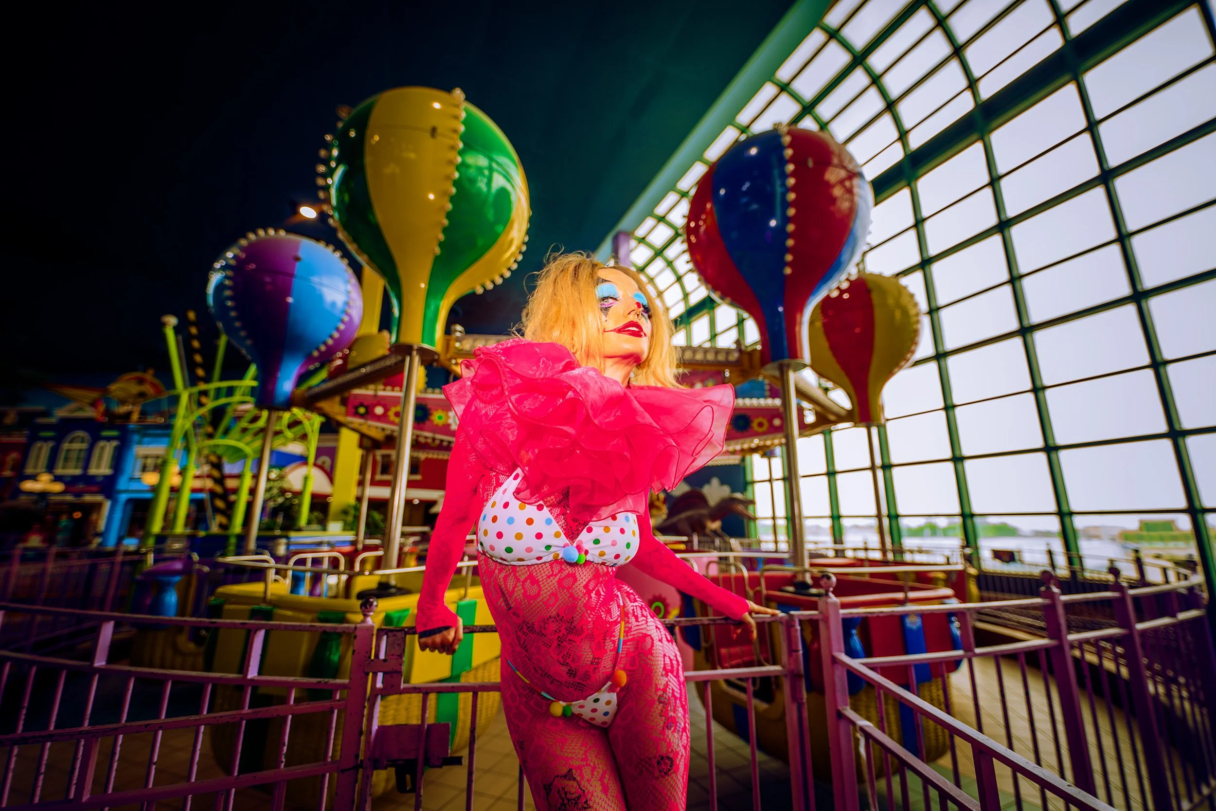 A clown dressed in bright pink with colorful makeup, standing in front of a carnival ride with colorful balloon-shaped decorations.