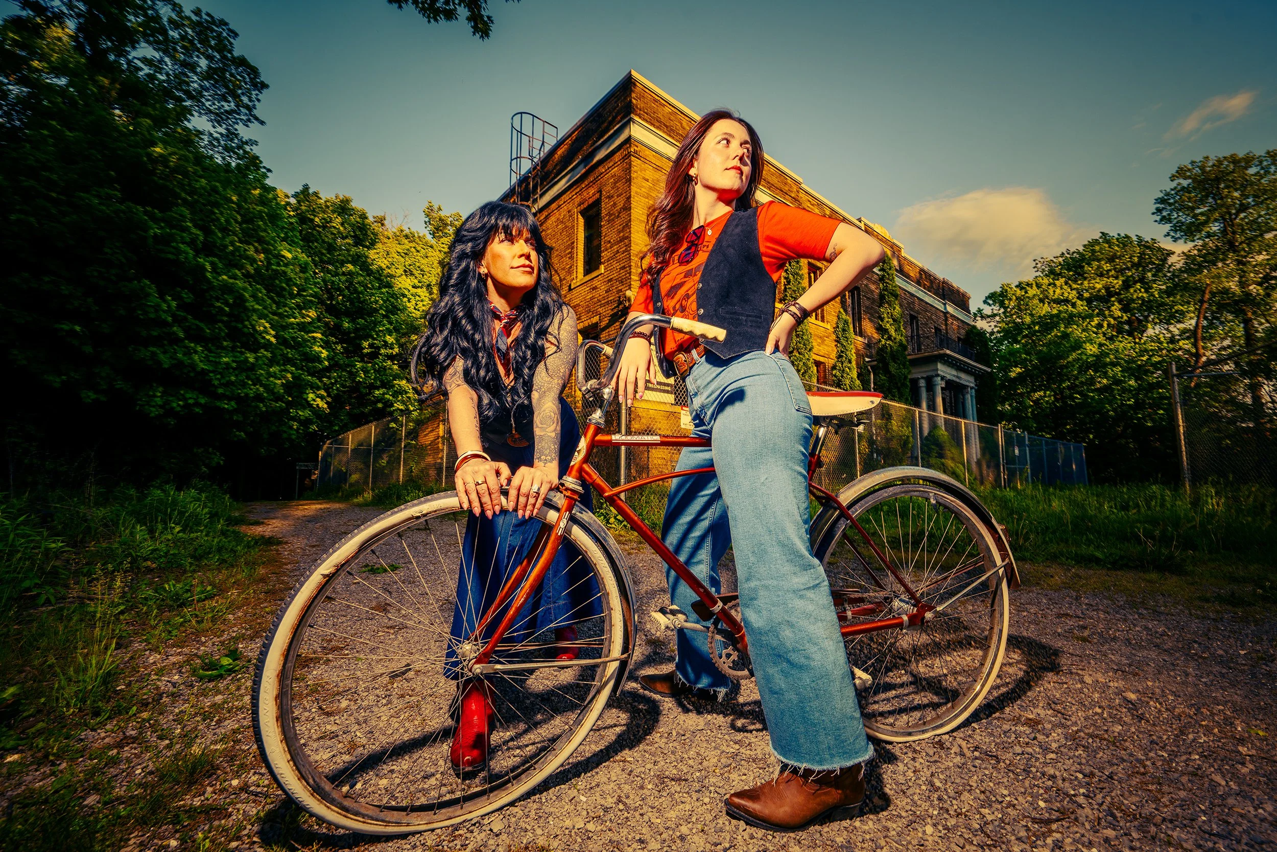 Two women standing beside a bicycle outdoors with trees and a brick building in the background during sunset or late afternoon.