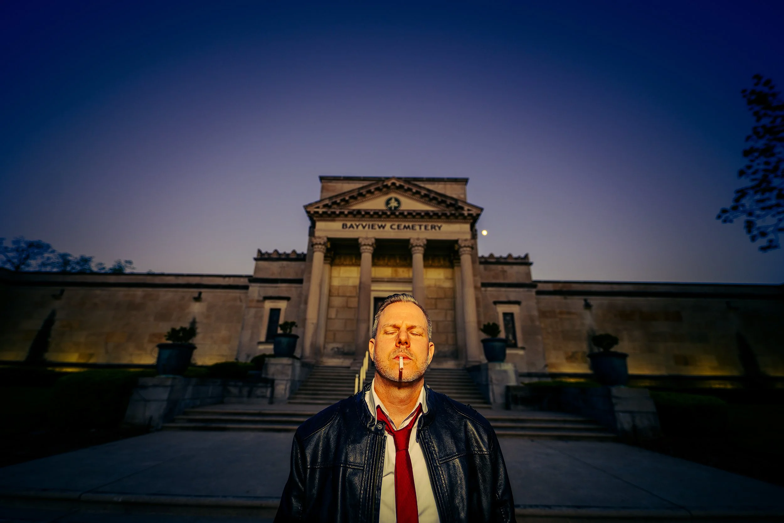 A man with eyes closed and a cigarette in his mouth standing in front of a historic courthouse building at dusk. The courthouse has a classical architectural style with columns and a sign that reads 'Bayview Cemetery.'