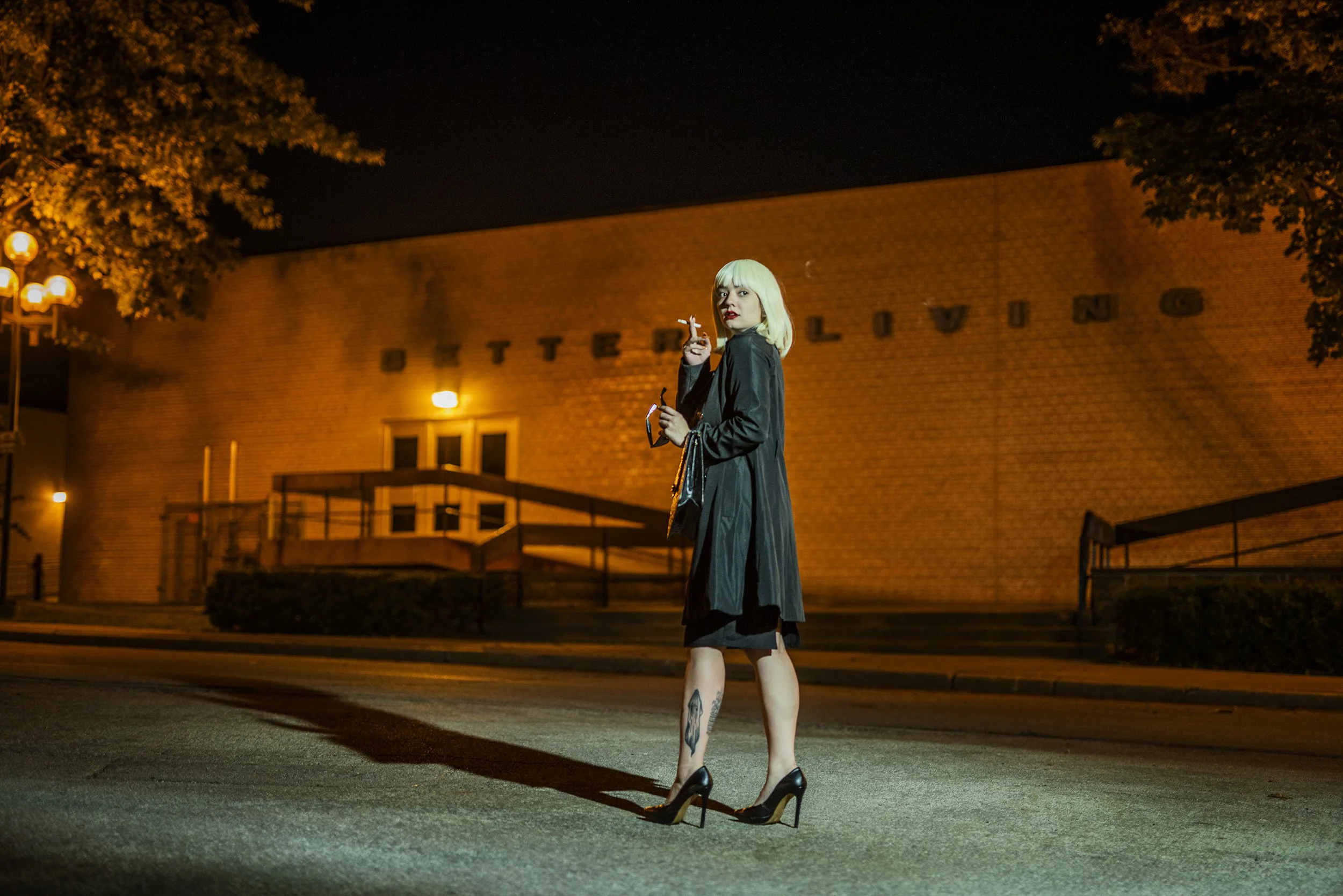 A woman with blond hair and tattoos on her legs standing outside at night in front of a brick building with the word 'BATTLE' on it. She is wearing a black coat, high heels, and holding a cigarette. Her high heels are on the ground while her feet res