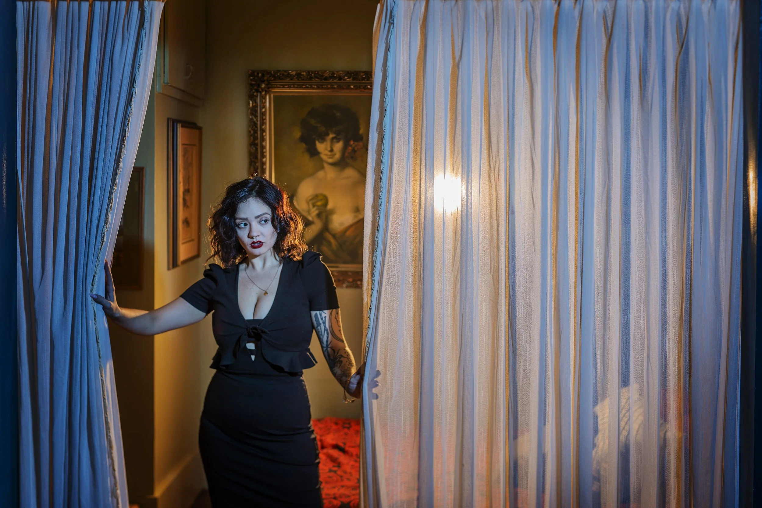A woman in a black dress with tattoos on her arm is standing in a room with yellow walls and floral carpet, holding a sheer curtain open. Behind her is framed artwork of a woman with dark hair, holding a yellow object.