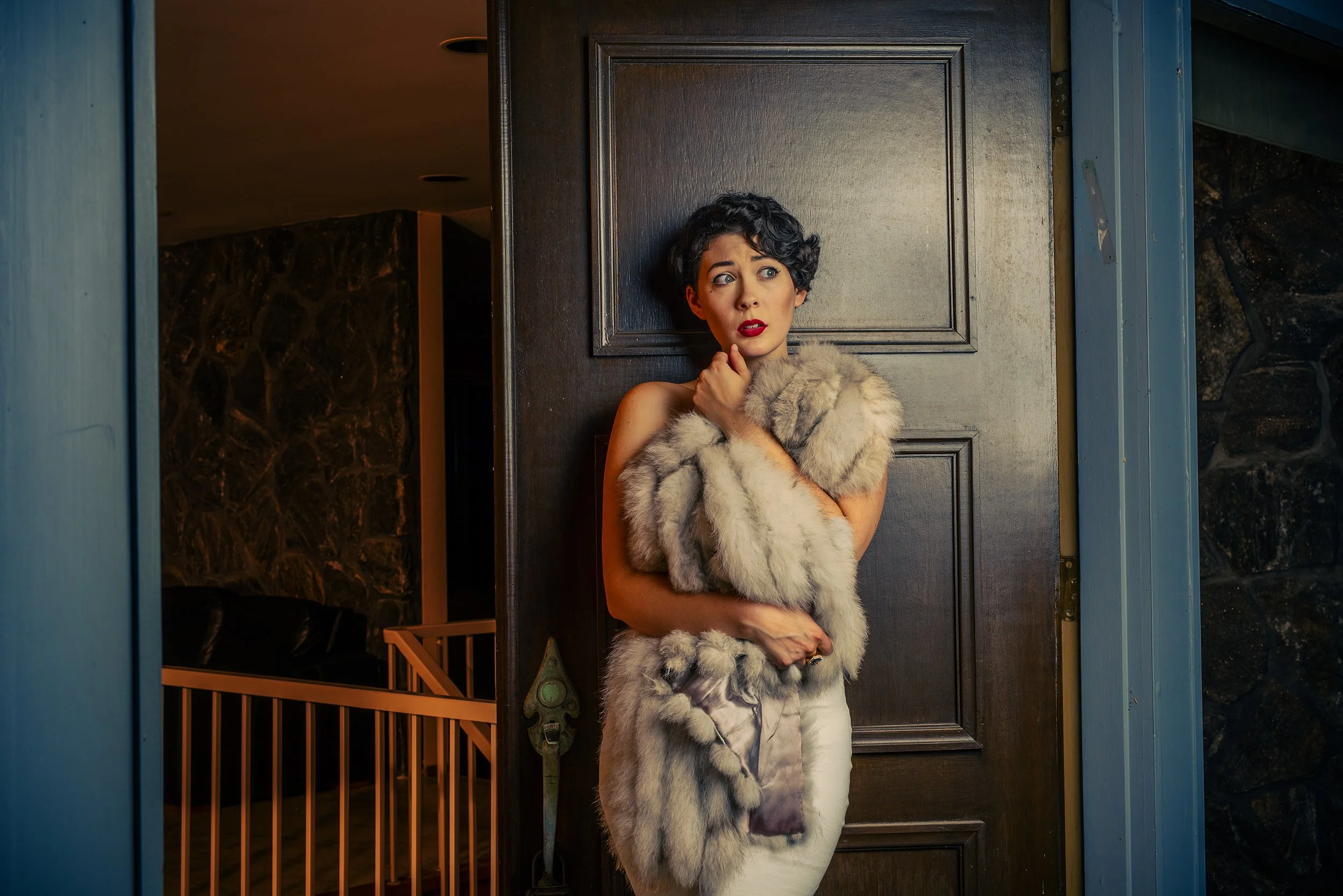 A woman with short curly black hair and red lipstick, wearing a beige fur stole, stands in a doorway looking to her left with a nervous expression.