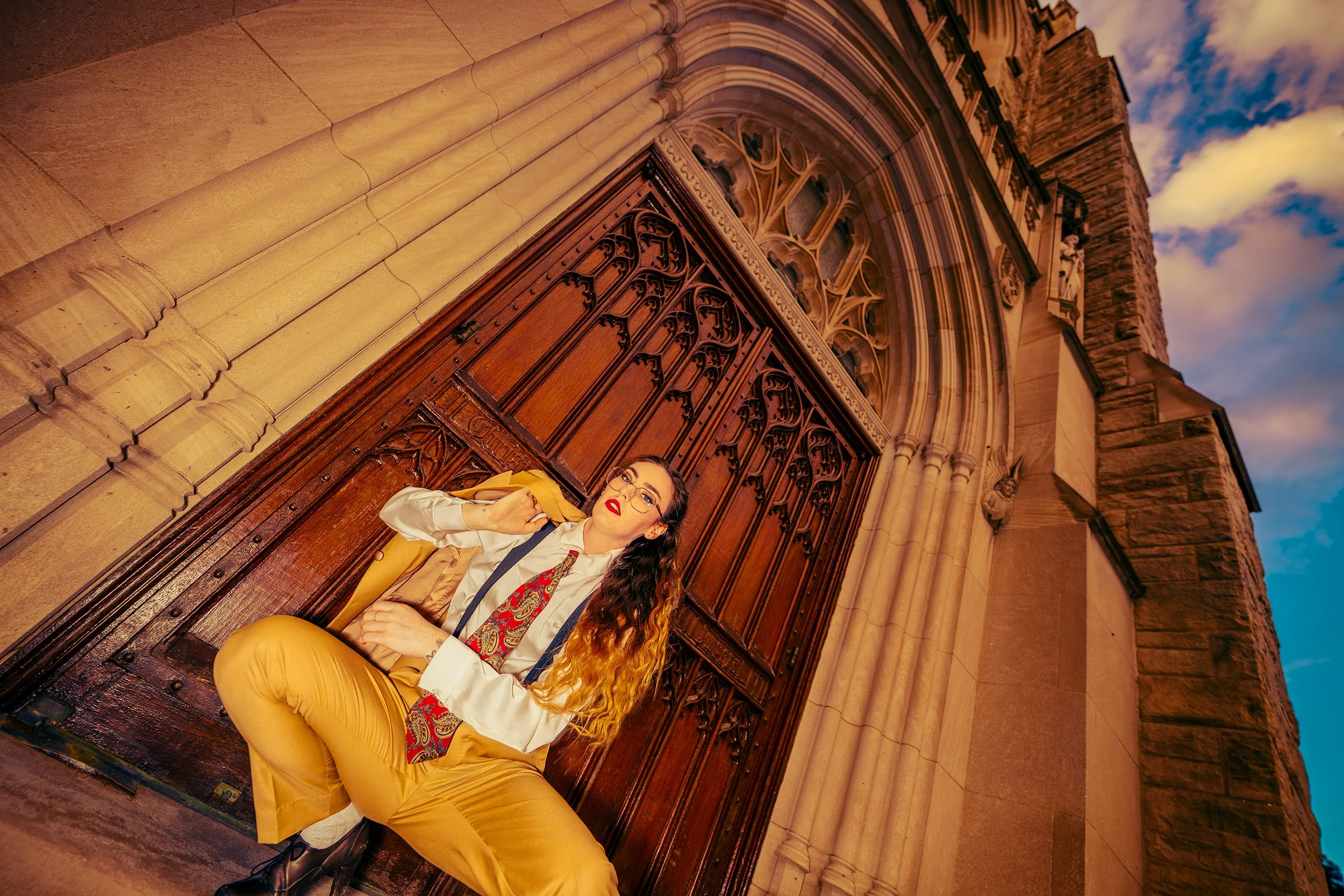 A woman with long, curly red hair, wearing glasses, a white shirt, a colorful scarf, and yellow pants, sitting on a wooden bench or door outside a historic stone building with gothic architecture and a blue sky with clouds in the background.