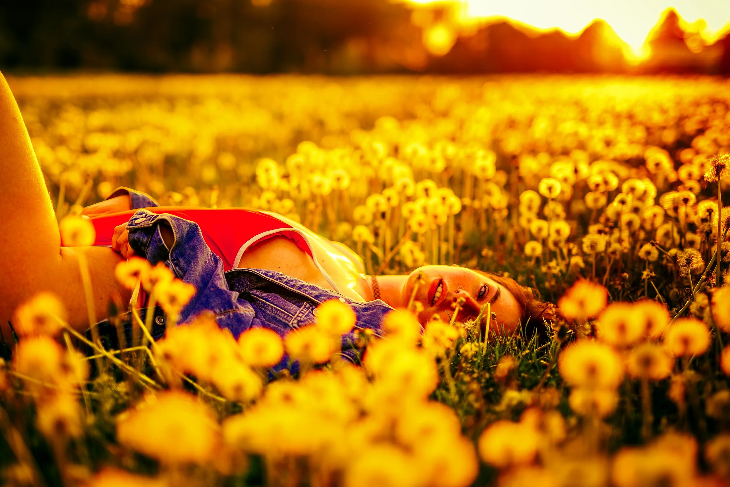 A person lying in a field of yellow flowers during sunset, smiling with eyes closed, wearing a denim jacket and red top.