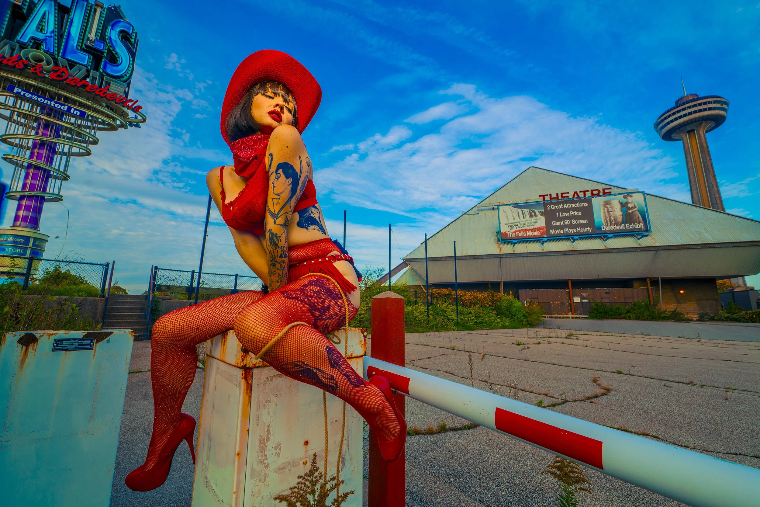 A woman with tattoos and red clothing, wearing a red hat, sitting on a rusted barrier outside an abandoned theater with a large billboard, a tall observation tower, and a sign that reads 'Theater' in the background on a partly cloudy sky day.