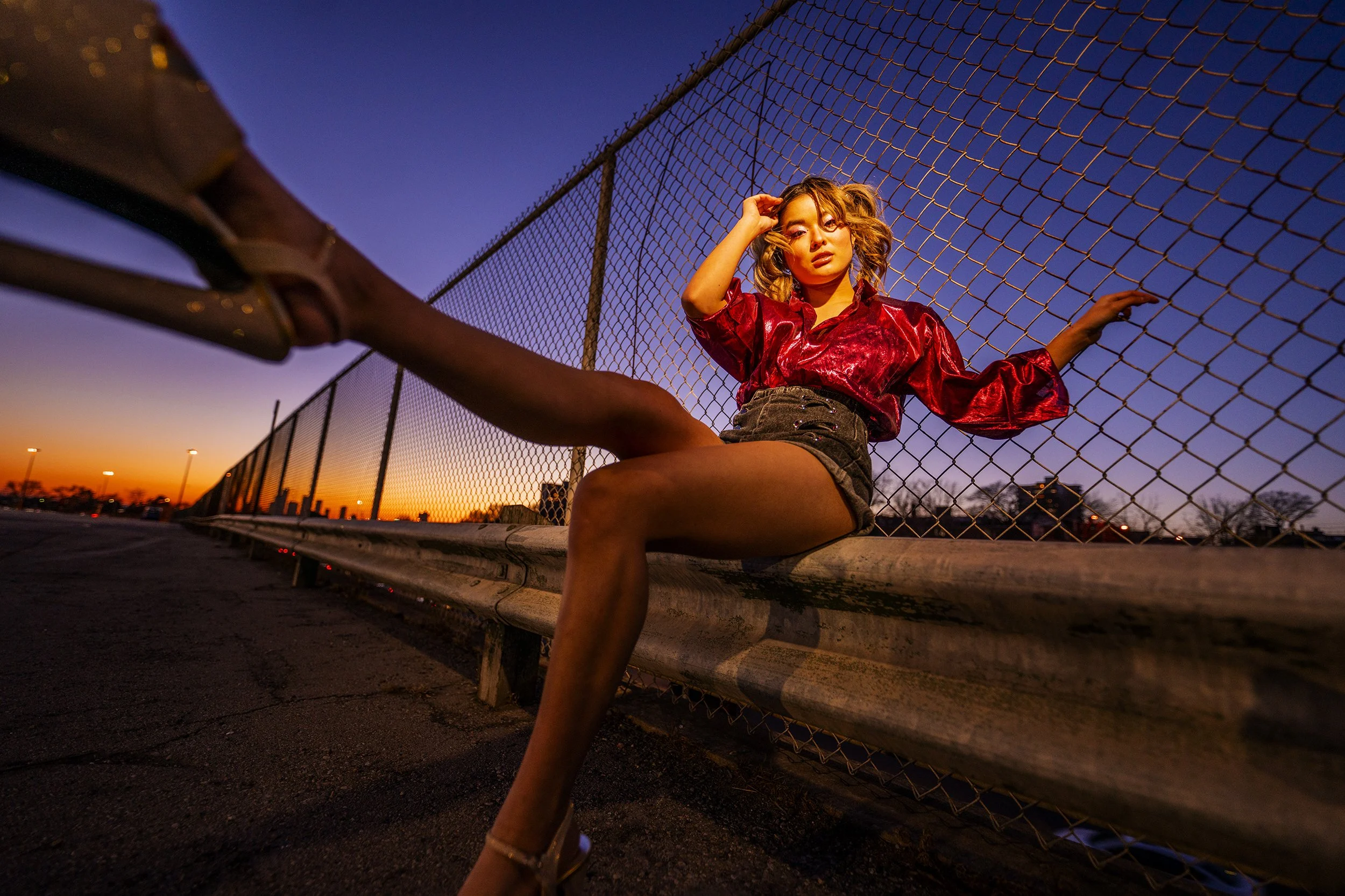 A young woman with curly hair wearing a shiny red top and dark shorts sitting on a concrete barrier in front of a chain-link fence at sunset.