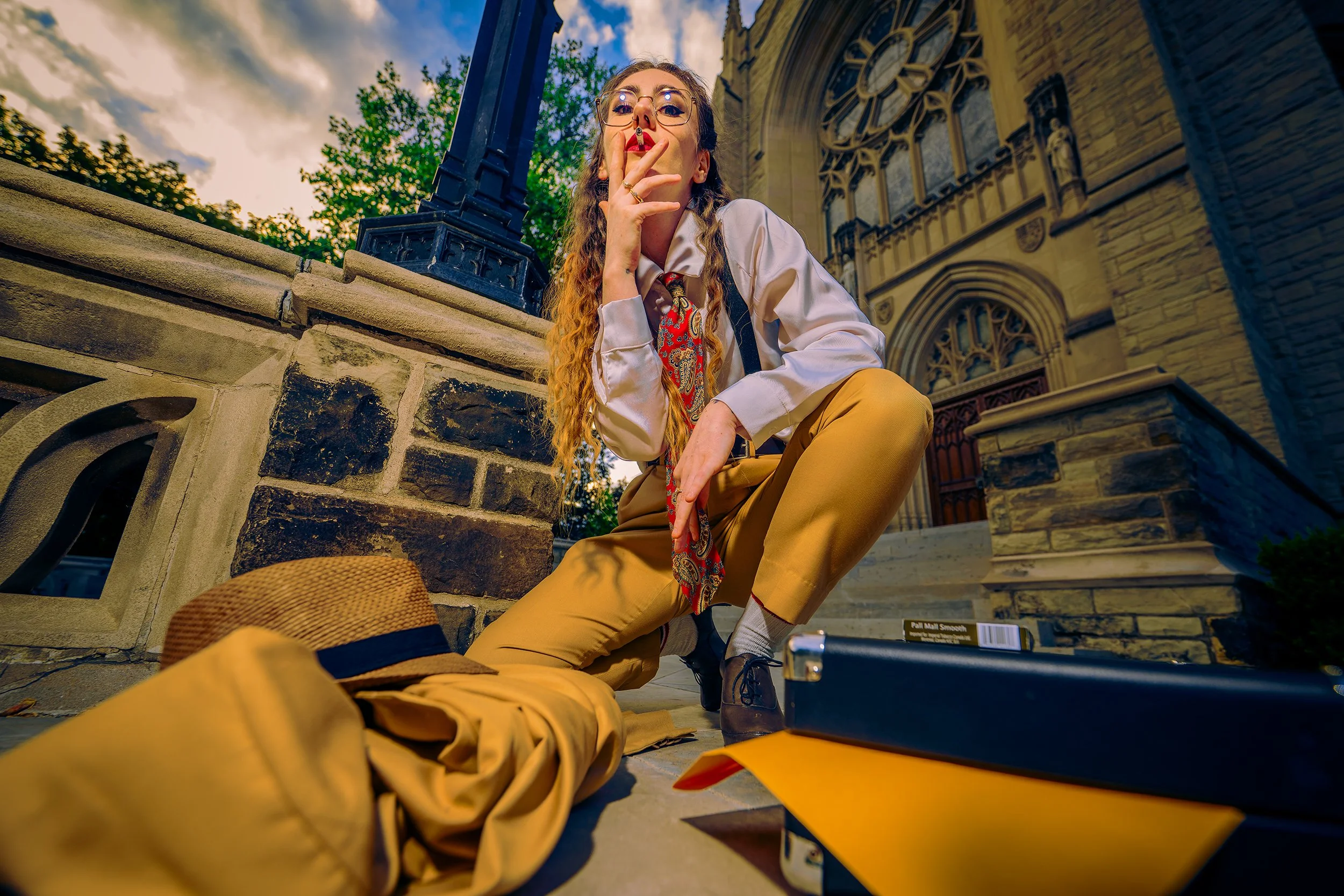 A woman with glasses and long curly hair smoking a cigarette while sitting on the steps of a historic building with castle-like architecture during sunset. She is wearing yellow pants, a white shirt, and a patterned tie, with a straw hat and a camera