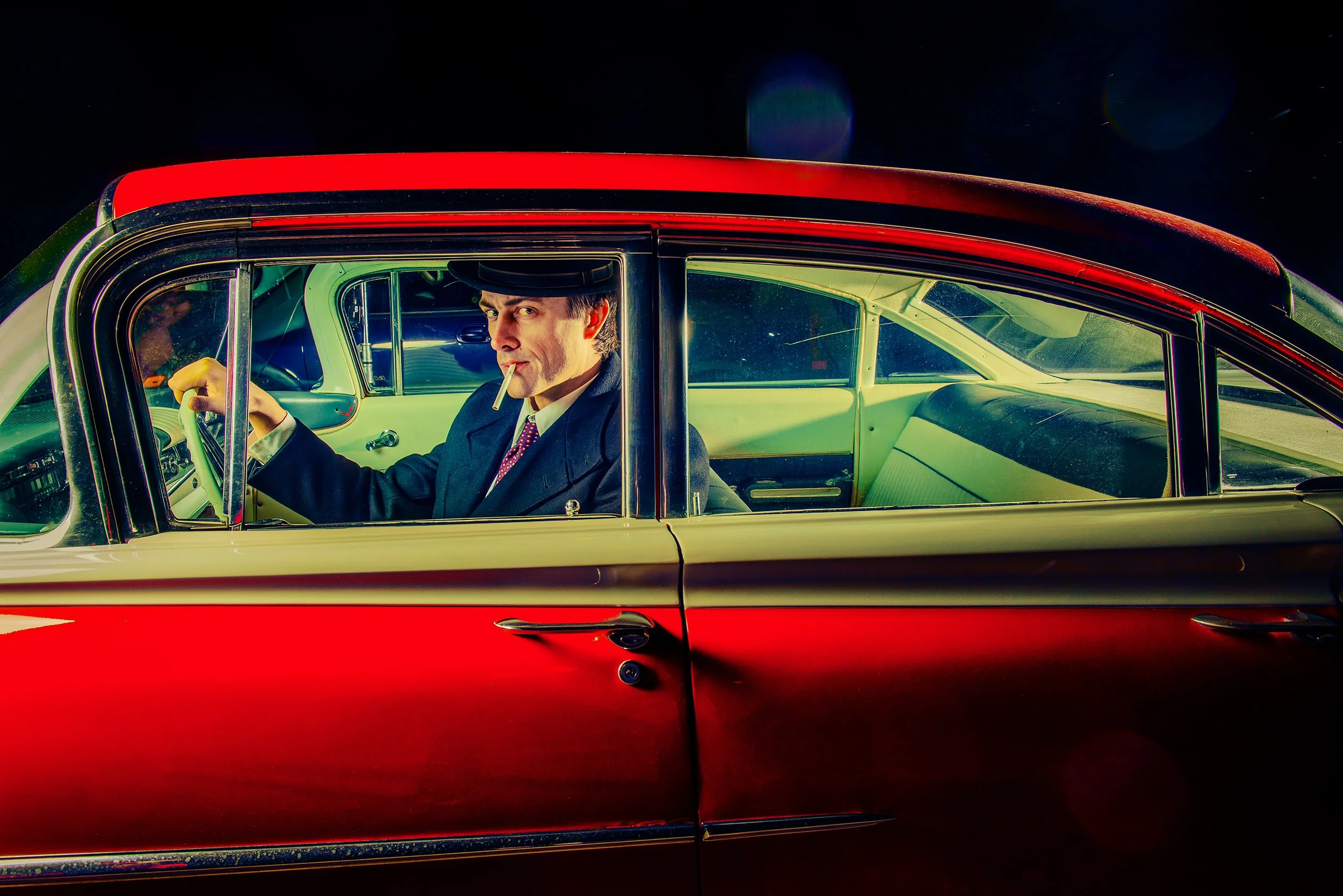 A man in vintage clothing with a cigarette in his mouth sitting in a classic red and white car at night.