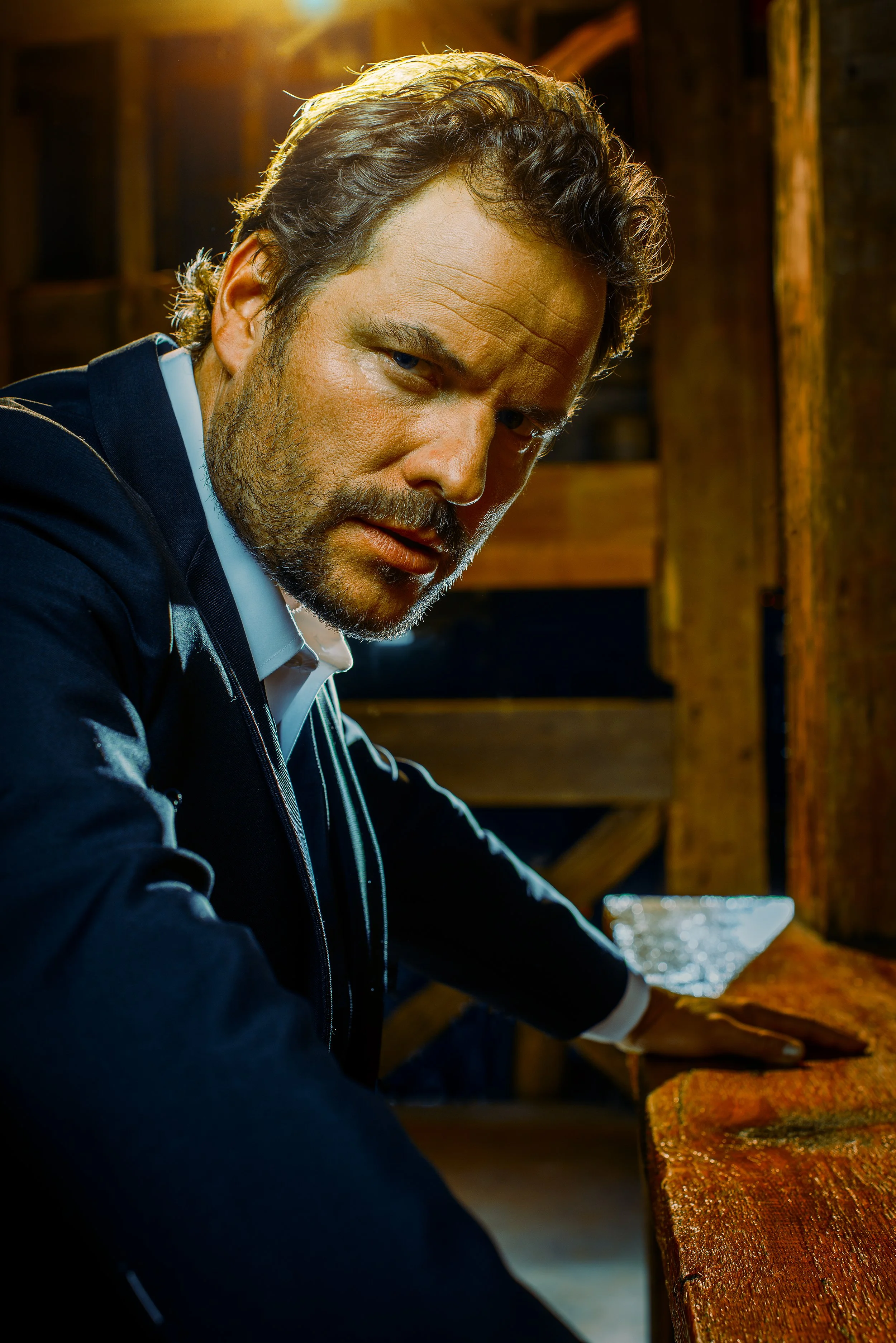A man with dark hair, beard, and mustache wearing a navy blazer and white shirt playing the piano in a dimly lit wooden room.