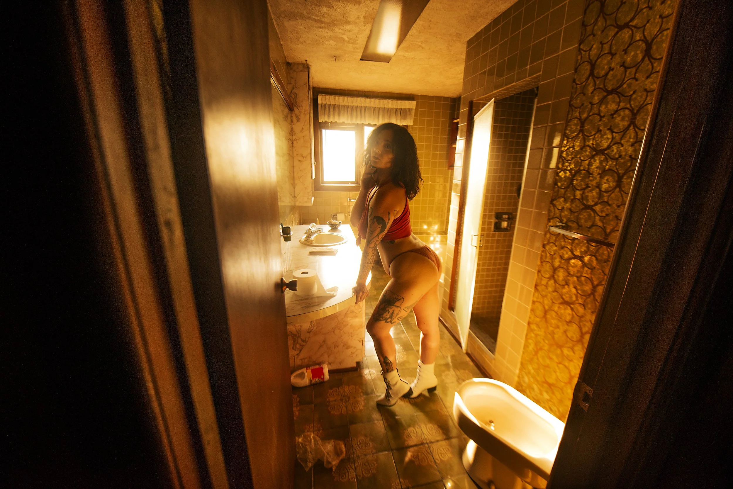 A woman with tattoos and wearing a red top, white socks, and orange shorts is standing in a vintage bathroom. She is looking towards the camera with her hand near her face, and sunlight is coming through a window behind her. The bathroom has brown ti
