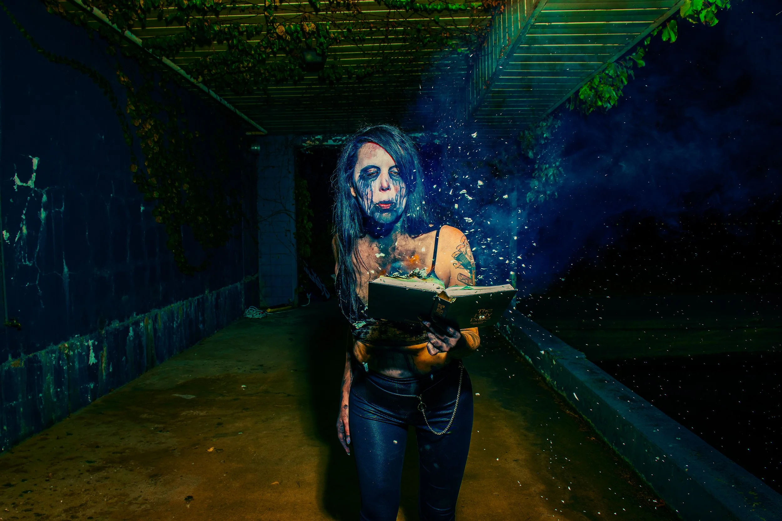 A woman with blue hair and face paint stands outside at night, reading a book and throwing some material into the air.