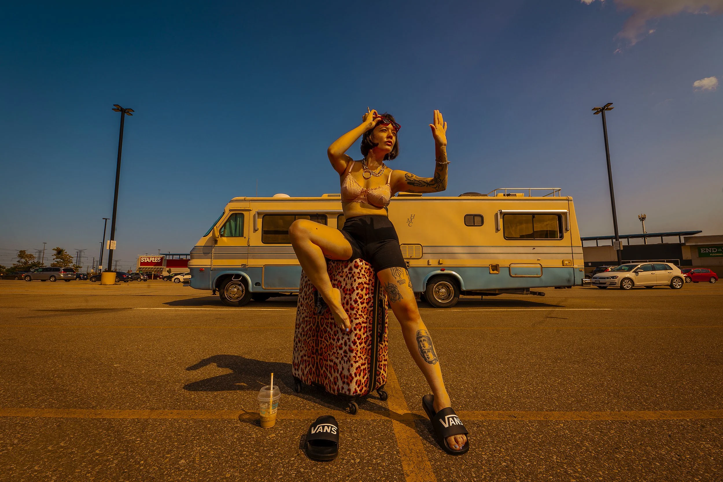 A young woman with tattoos, wearing a lace top, shorts, and slides, sits on a leopard print suitcase in a parking lot at sunset. She is posing with one knee up and arms raised, with an RV and several cars in the background.