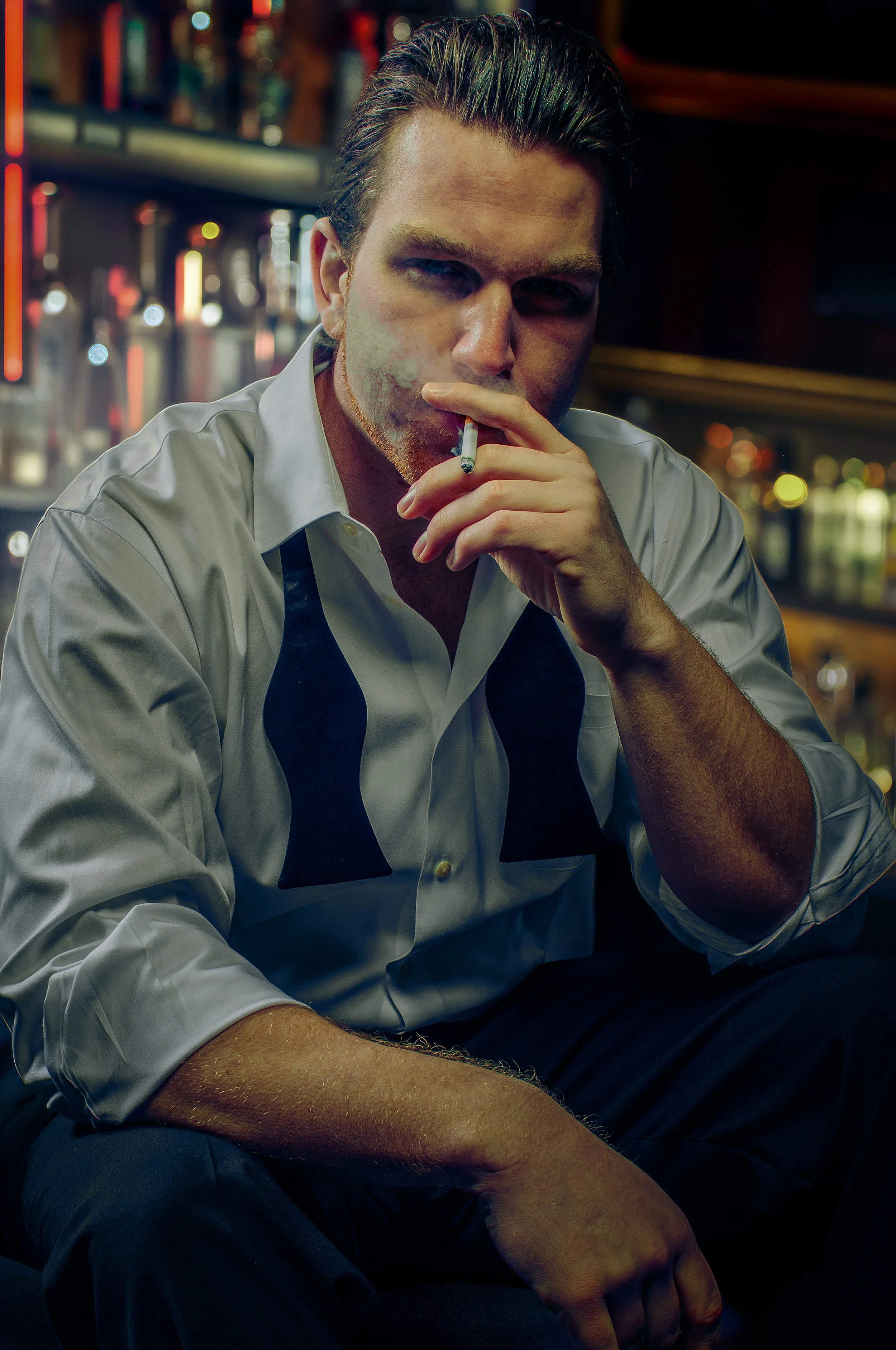 Man with slicked-back hair wearing a light dress shirt with a black tie, smoking a cigarette in a dimly lit bar with blurred colorful bottles in the background.