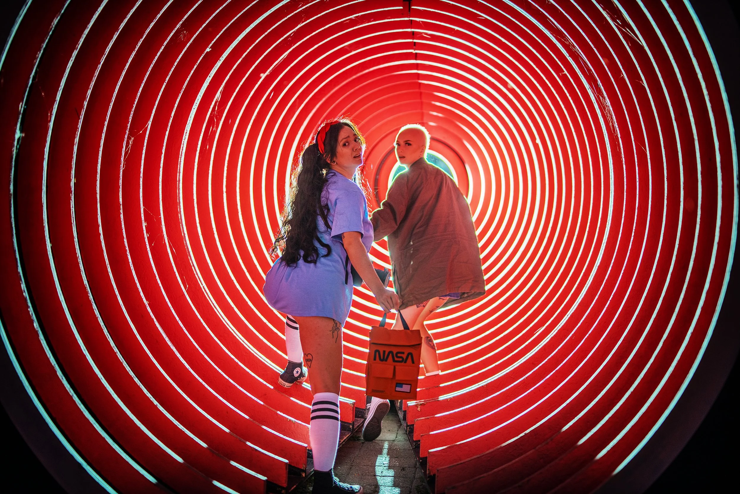 Two young women holding hands and walking through a red neon tunnel, one wearing a purple shirt and striped socks, the other in a brown jacket, with a NASA bag.
