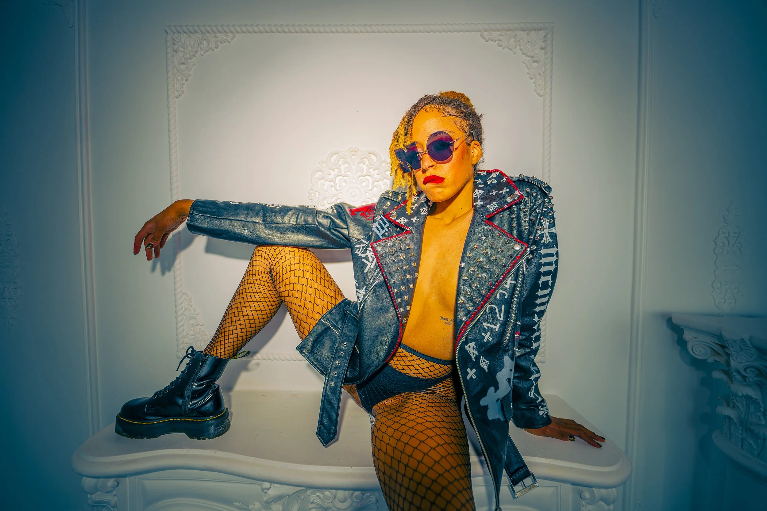 A person with blonde dreadlocks wearing dark sunglasses, a studded leather jacket, fishnet stockings, black shorts, and combat boots, striking a pose on a white ornate table in a decorated room with white walls.