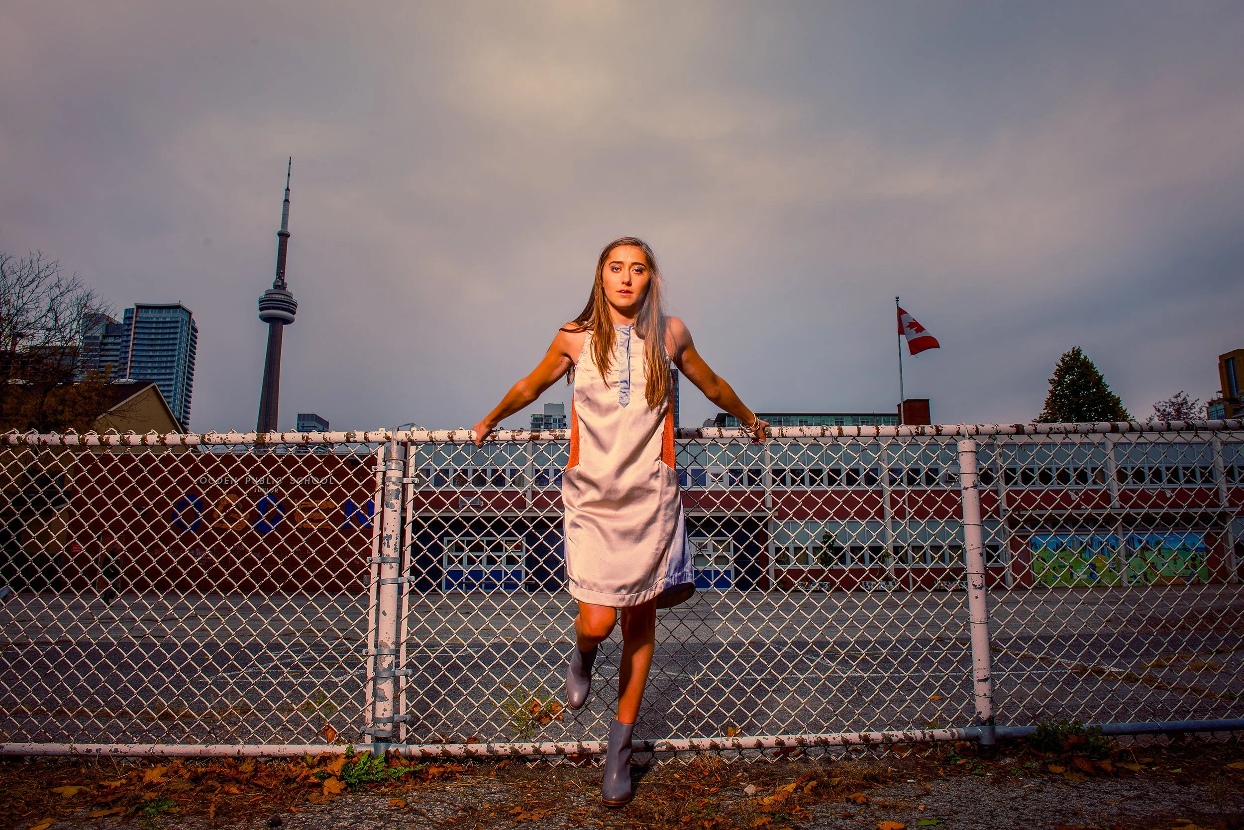 A young woman in a white dress with gray boots poses with her arms outstretched, holding onto a chain-link fence on a rooftop, with the CN Tower and Toronto skyline in the background during dusk.