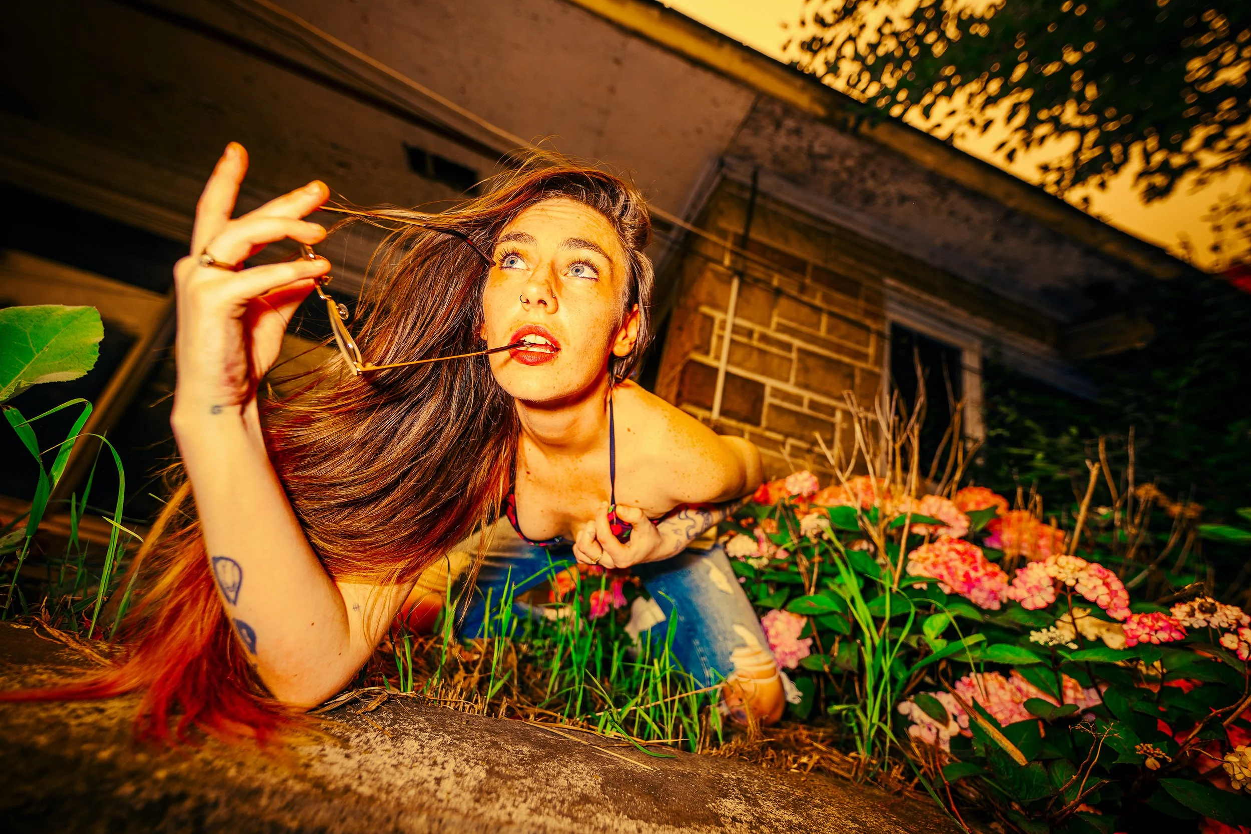 Young woman with long hair, tattoos, and makeup lying in a garden surrounded by pink and orange flowers, holding glasses in her hand, looking upward, in a backyard at sunset.