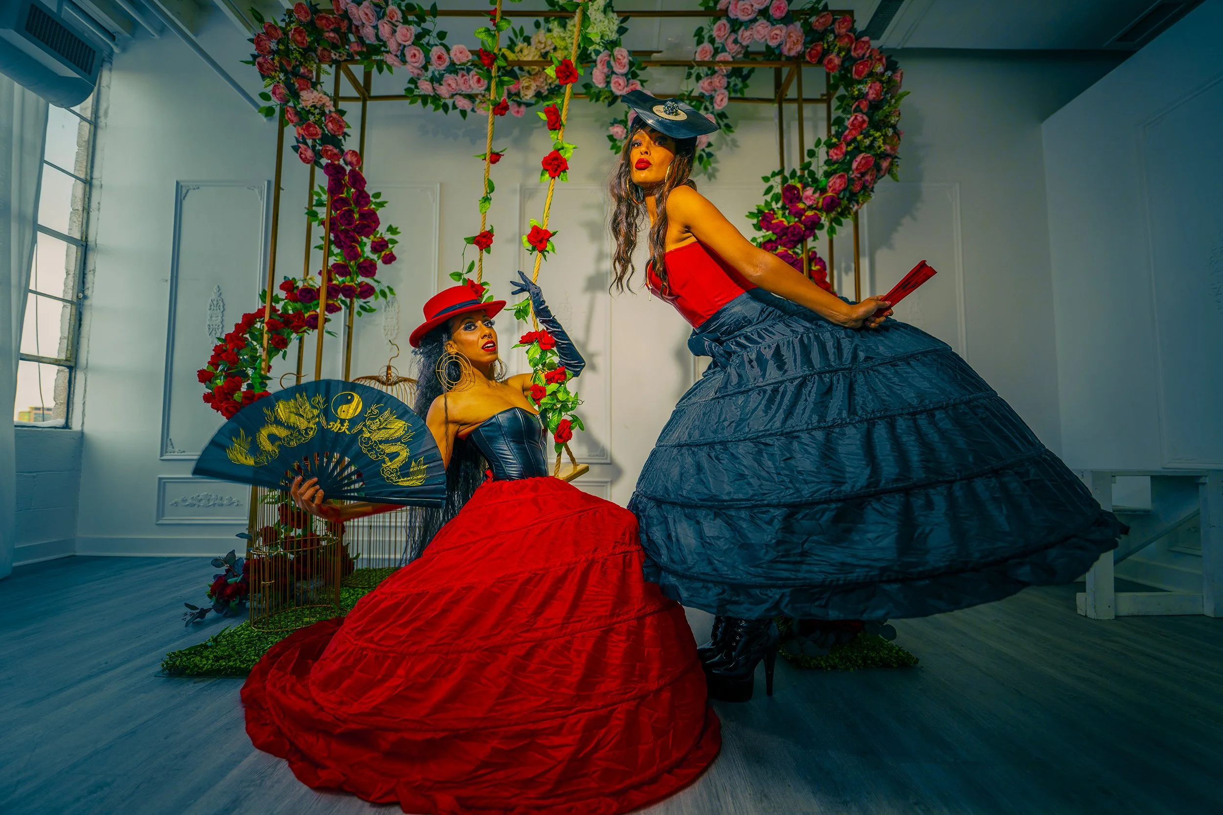 Two women dressed in elaborate costumes, holding fans and standing in front of a floral heart-shaped arch with a swing decorated with red roses, all within a decorated indoor setting.