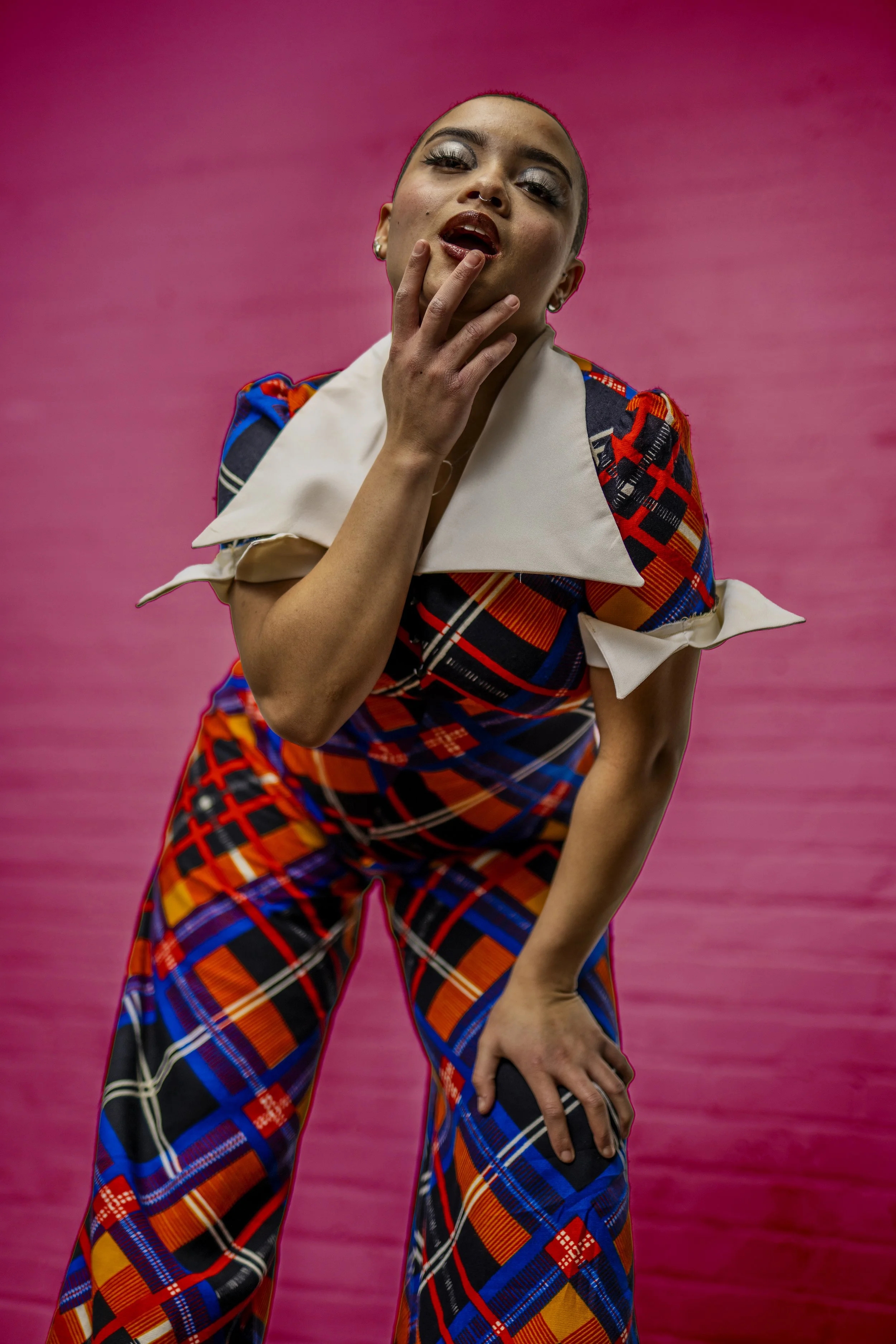 A person with short hair and makeup, wearing a colorful plaid outfit, poses with one hand on their hip and the other touching their face, against a pink background.