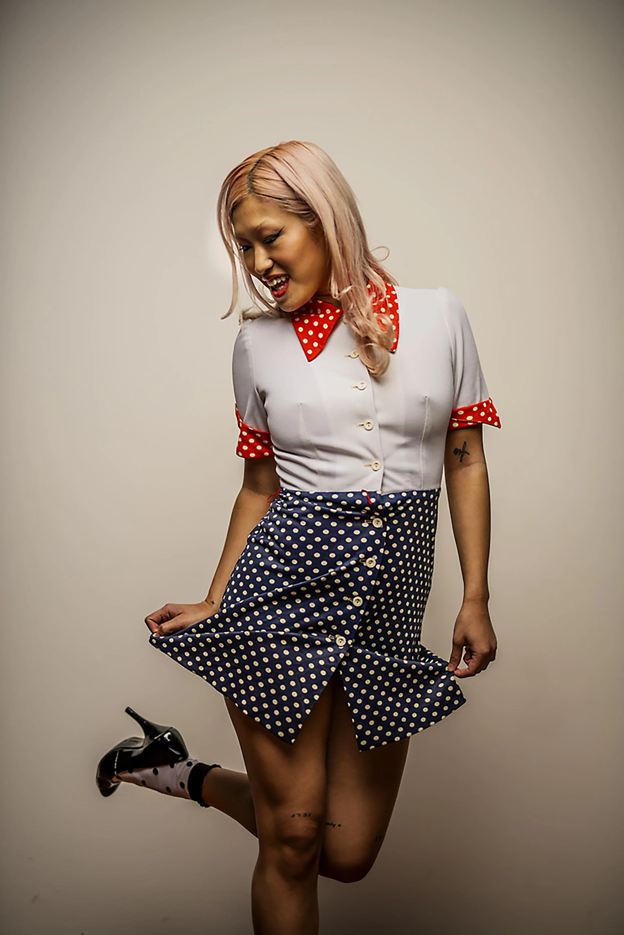A woman with pink hair wearing a vintage-inspired outfit featuring a polka-dot skirt and a white blouse with red polka-dot accents, posing with a playful expression against a plain background.