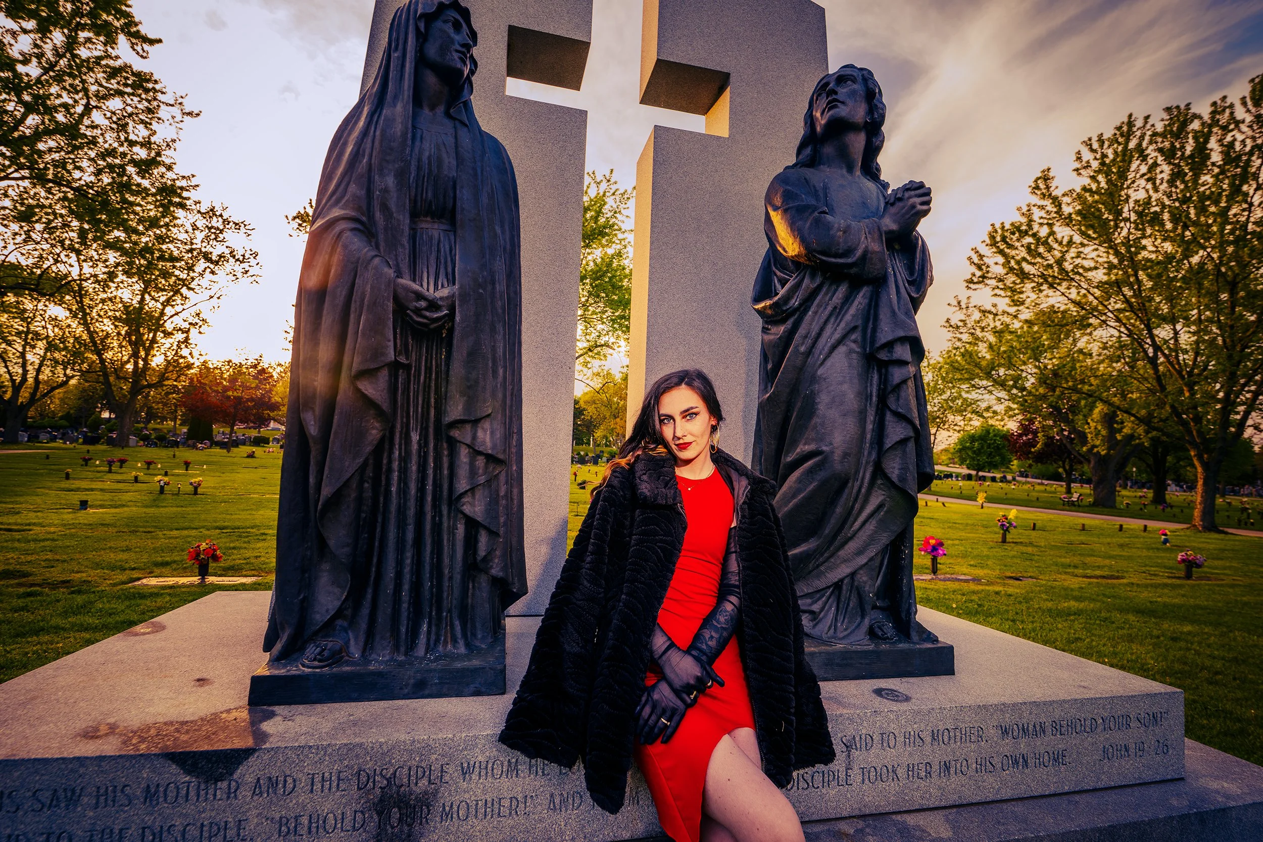 A woman with dark hair and red lipstick posing in front of a memorial with two bronze statues of women, one with hands clasped, and a large cross in the background during sunset in a cemetery surrounded by trees and flowers.