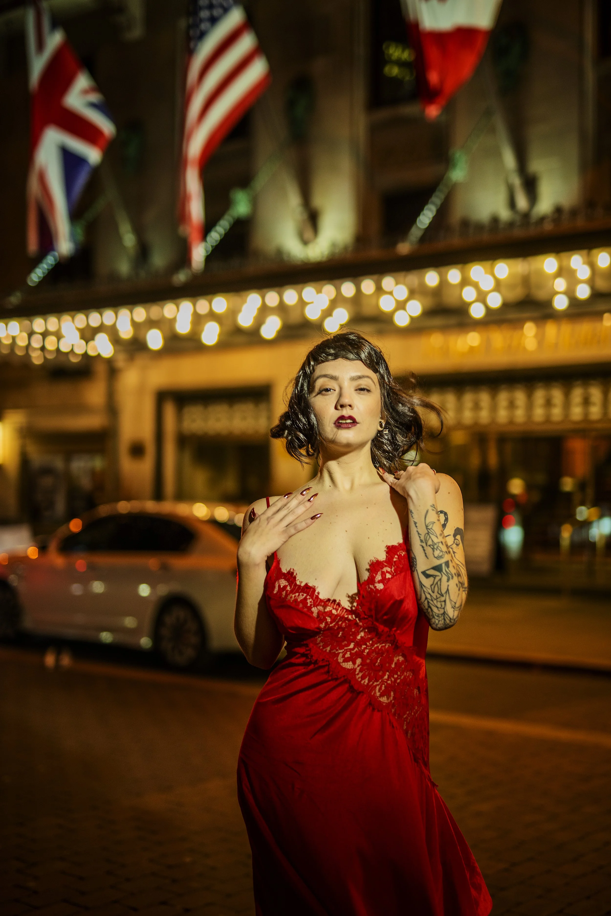 A woman with dark hair in a red dress standing on a city street at night, with flags and string lights in the background.