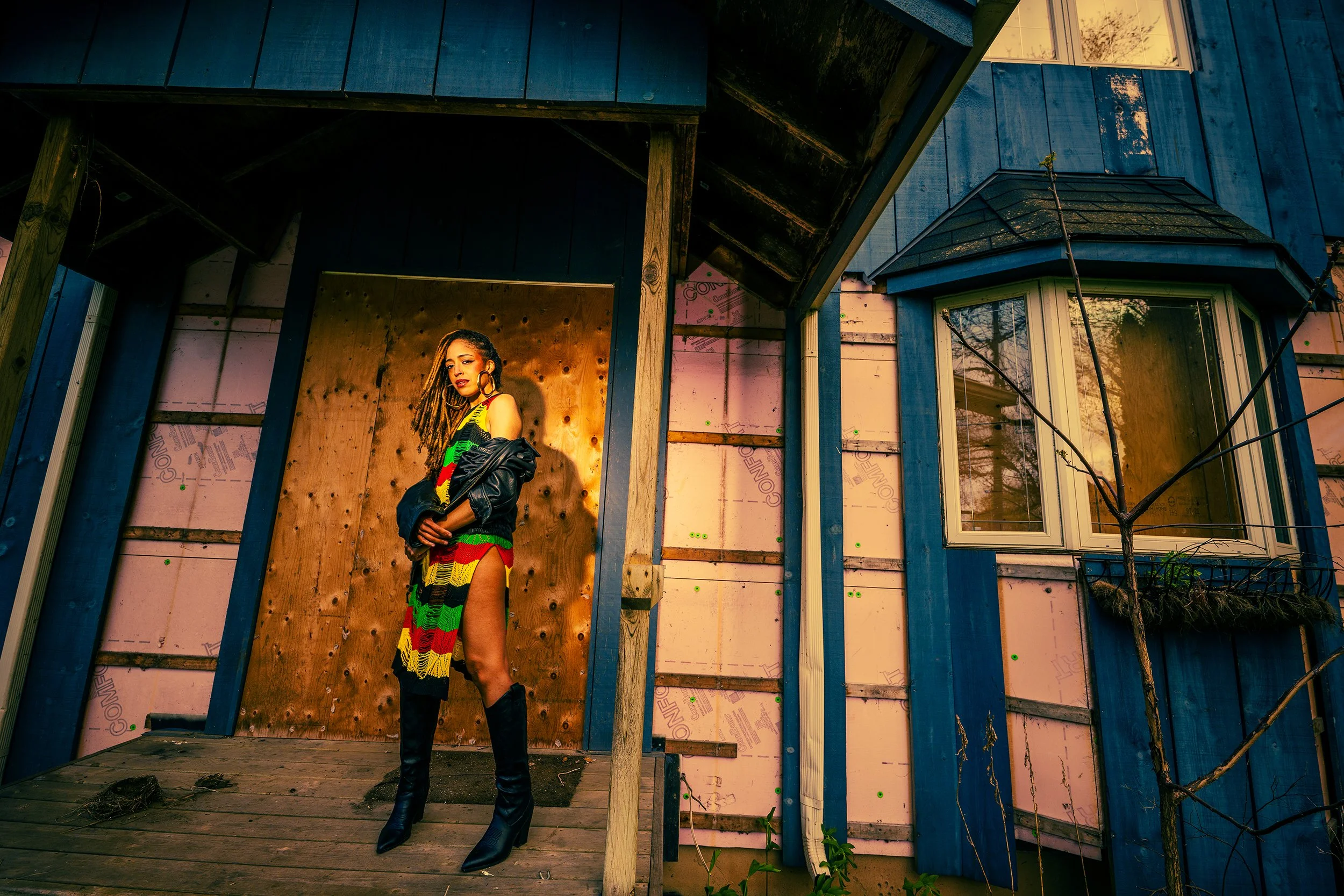 A woman with dreadlocks wearing a colorful, striped dress and black boots poses on a wooden porch in front of a partially constructed or renovated house with blue siding and window.