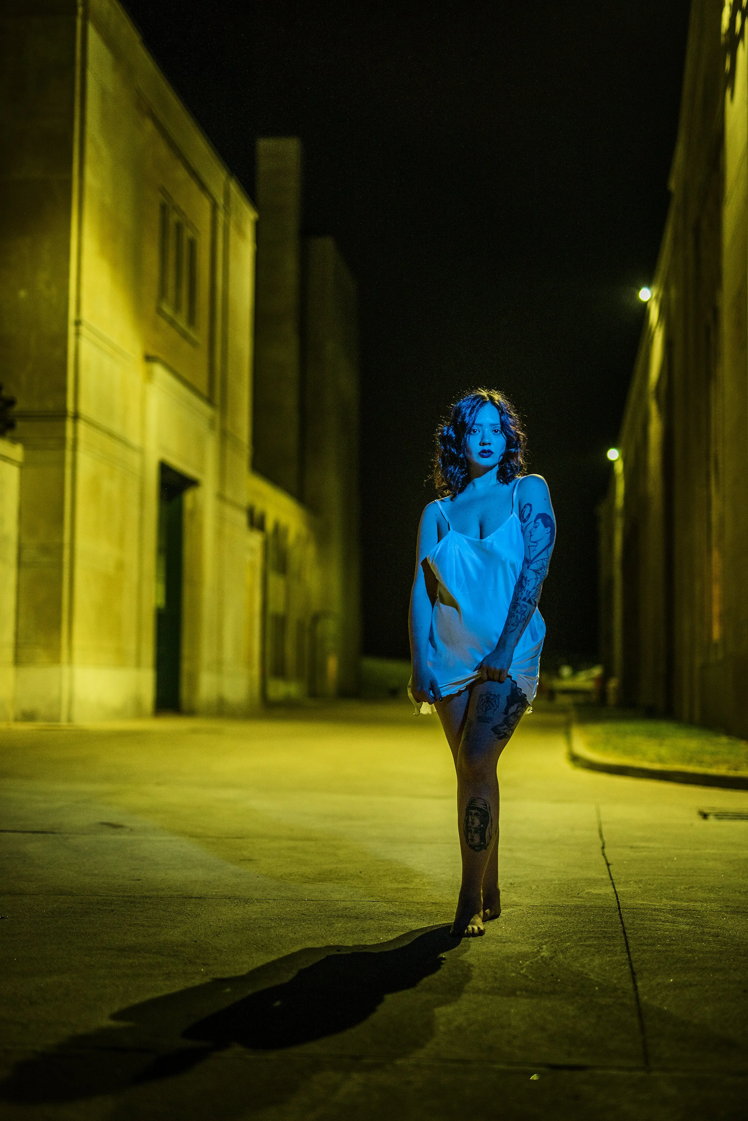 A woman with tattoos and curly hair walking on an empty street at night, illuminated by yellow and green streetlights, casting a shadow on the ground.