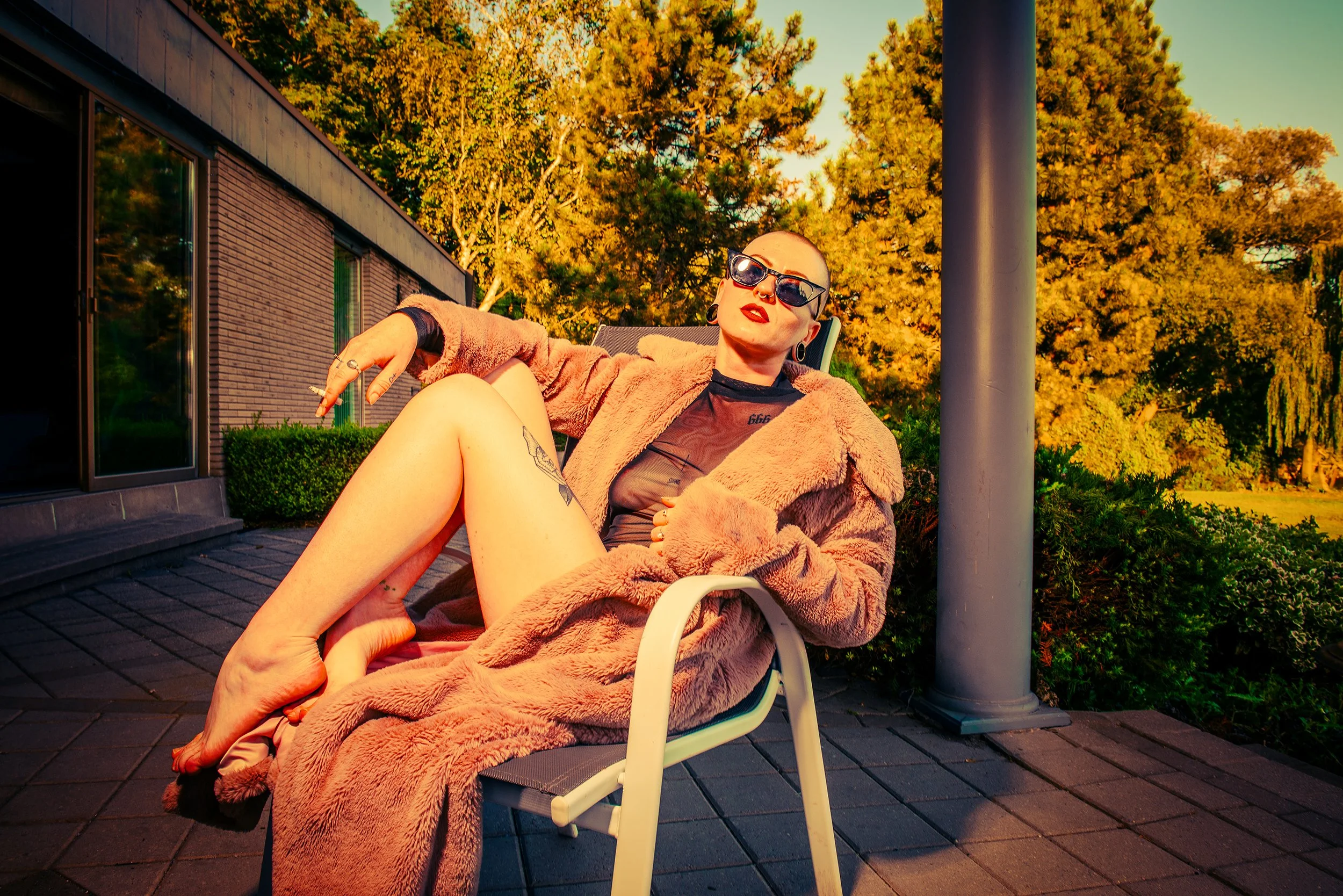 A person with a shaved head, sunglasses, and tattoos sitting on a lounge chair outdoors during sunset, wearing a fur coat, with a background of autumn-colored trees and a building.