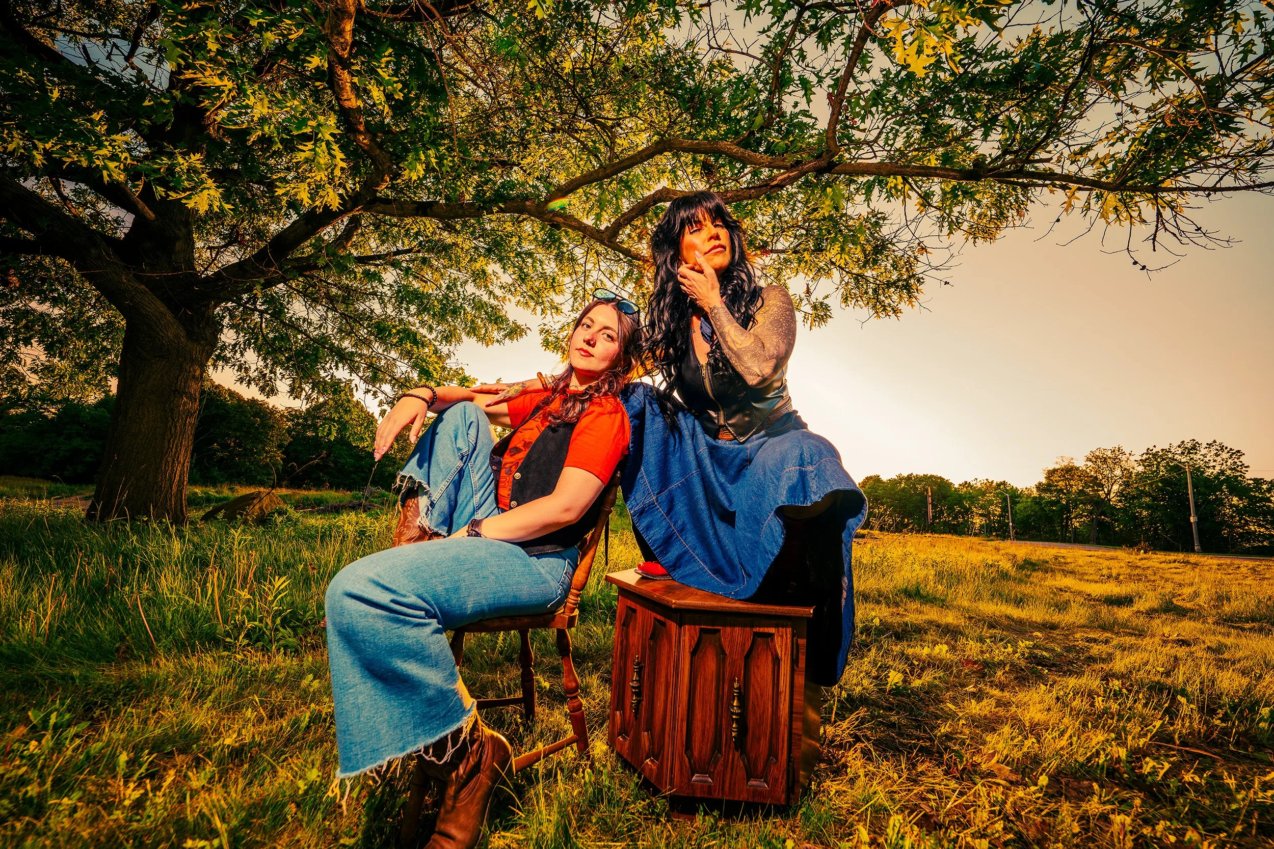 Two women sitting outdoors beneath a large tree during sunset, with one woman leaning on a small wooden table and the other touching her face, both looking contemplative.