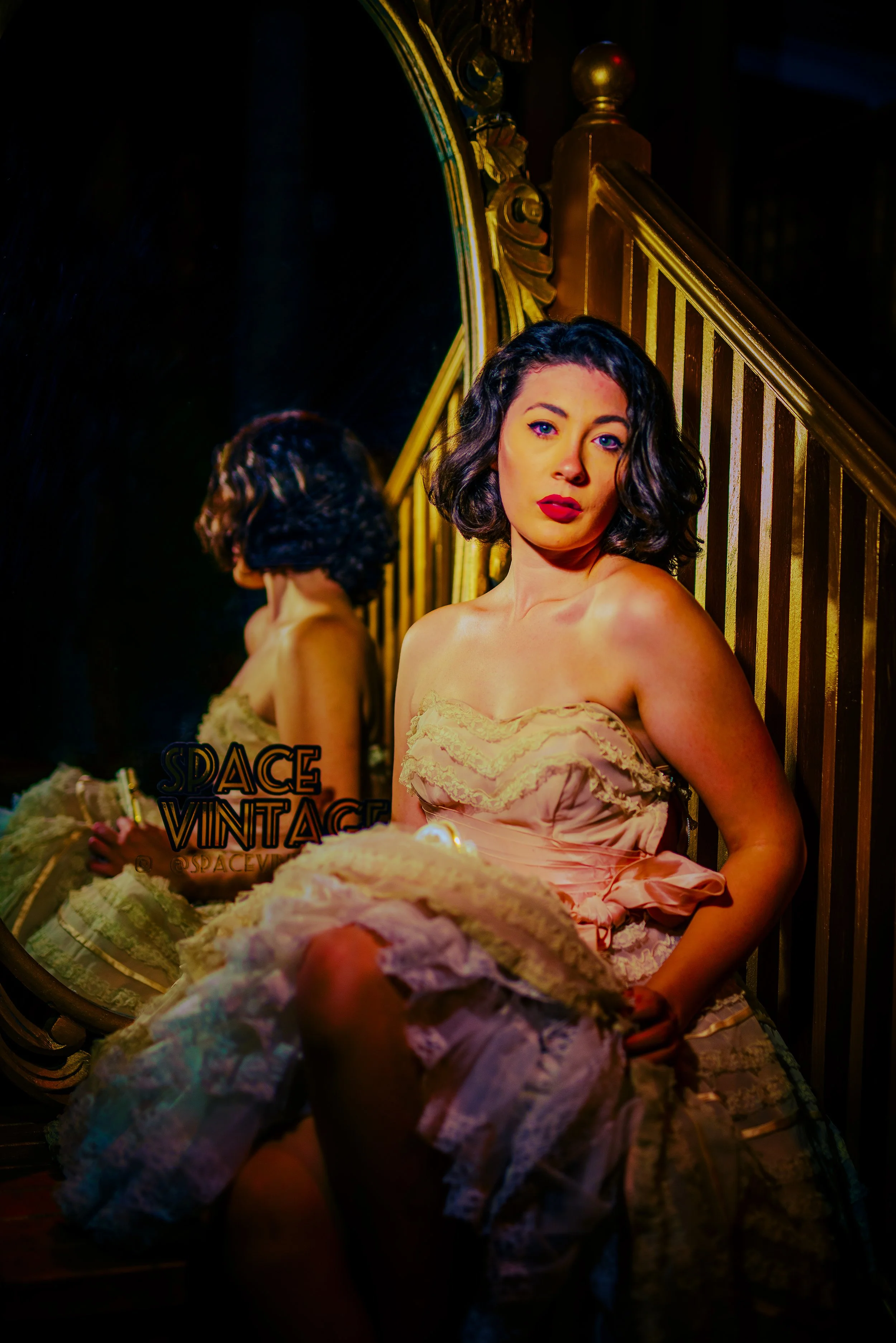 A woman with short curly dark hair and blue eyes, wearing a pink satin dress, sitting by a gold-trimmed mirror with a gold staircase in the background. The mirror reflects her profile and part of a woman in a vintage dress with floral patterns. The s