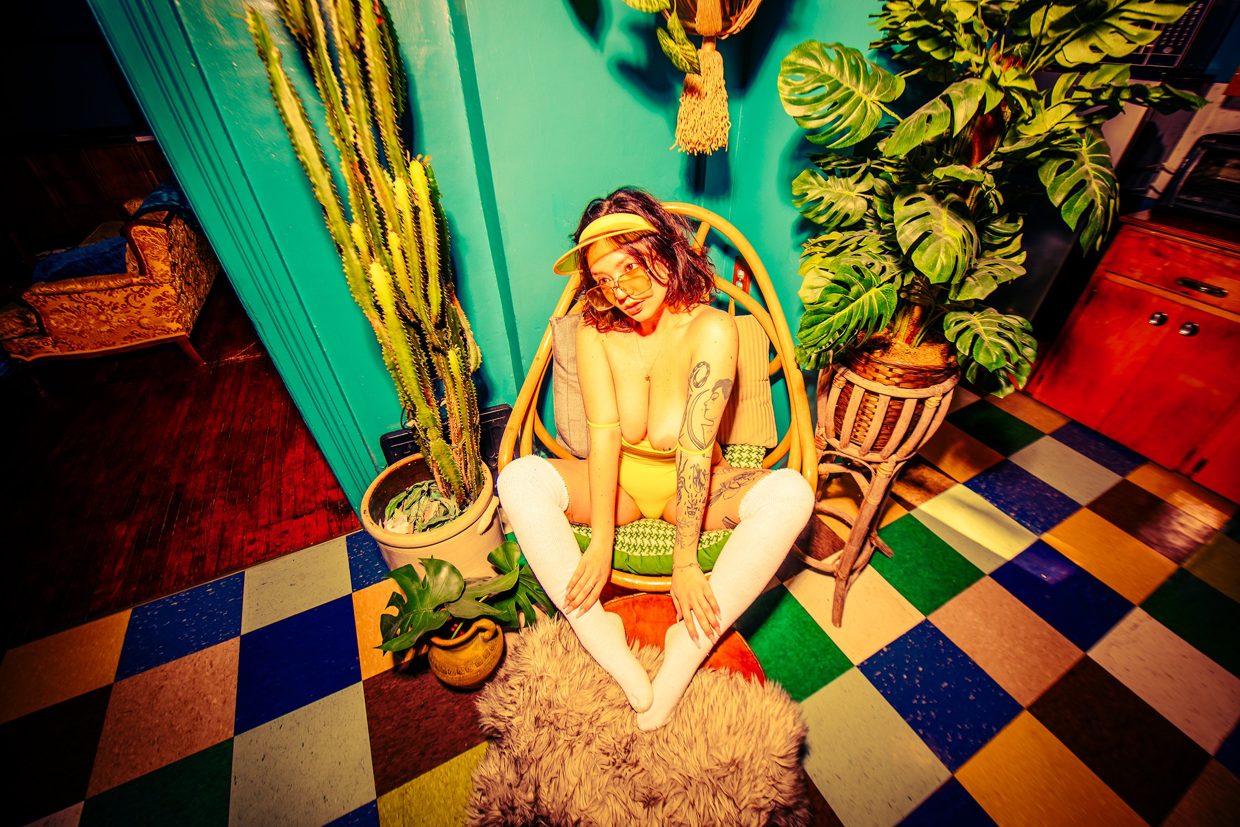 A woman with glasses and tattoos sitting on a wooden chair surrounded by potted plants in a colorful, eclectic room with tiled flooring.
