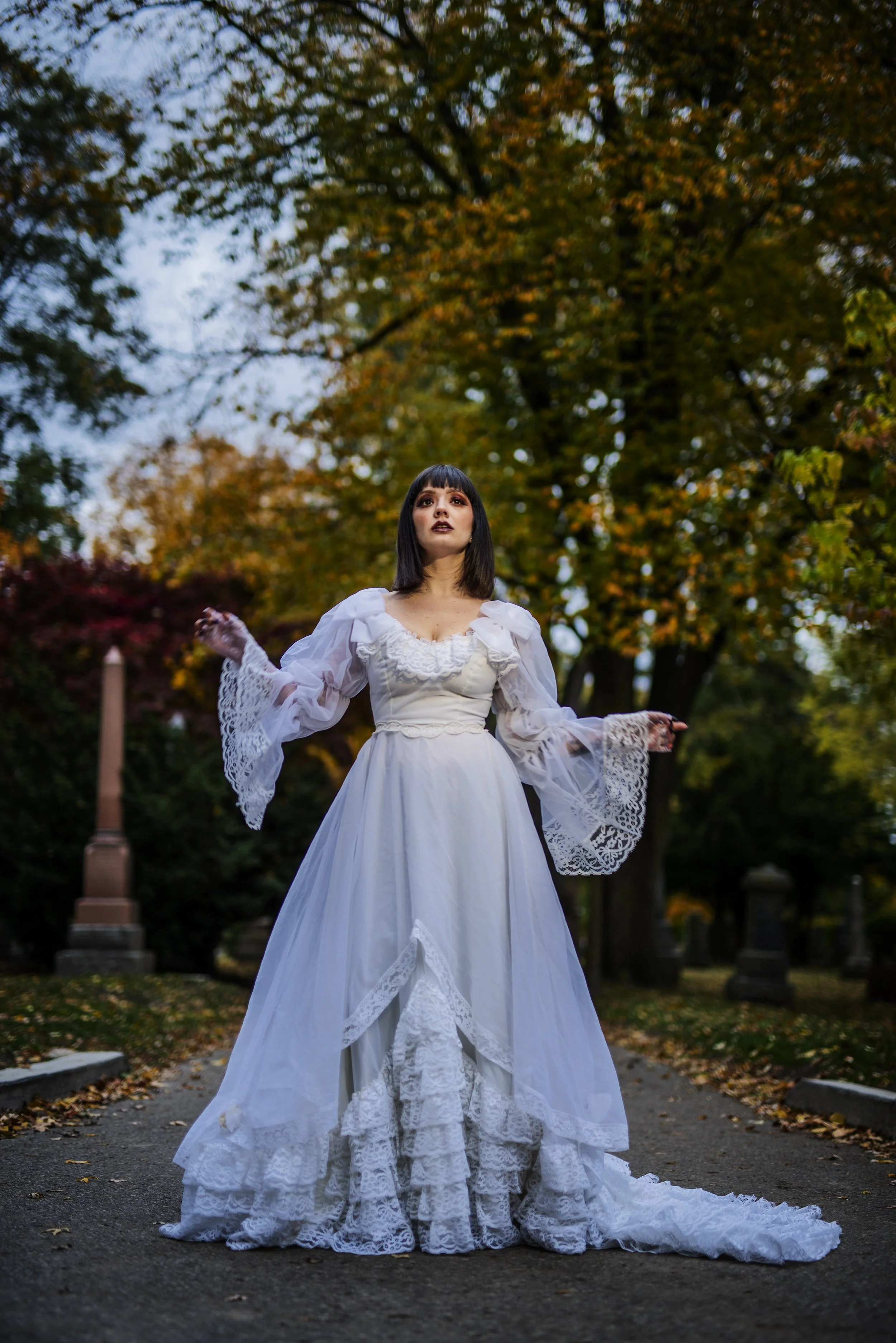 A woman dressed in a vintage style white wedding gown with lace details, standing outdoors in a cemetery with trees displaying autumn leaves.