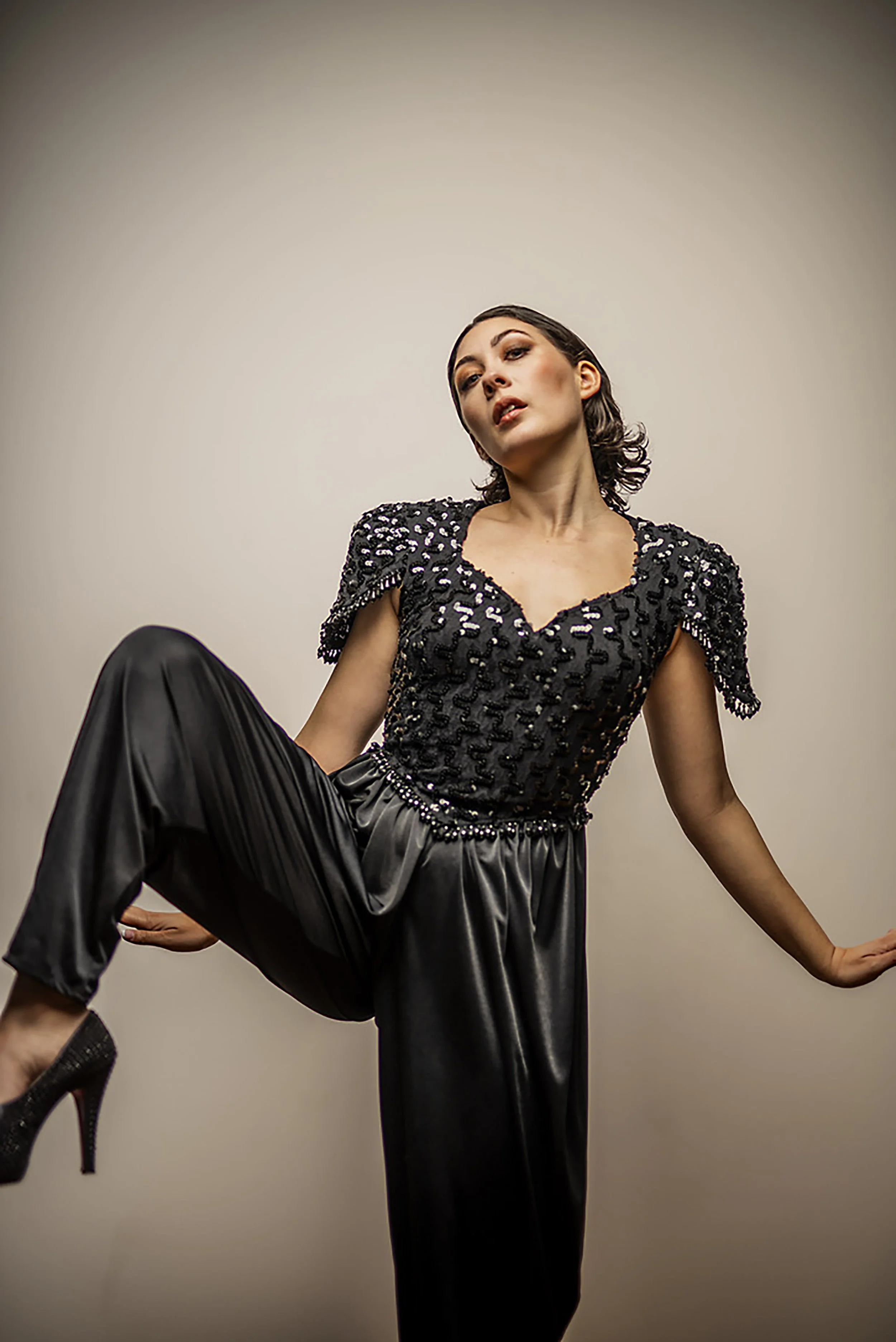 A woman with short dark hair and makeup wearing a black sequined top and black pants, posing with one leg raised and arms extended against a plain light-colored background.