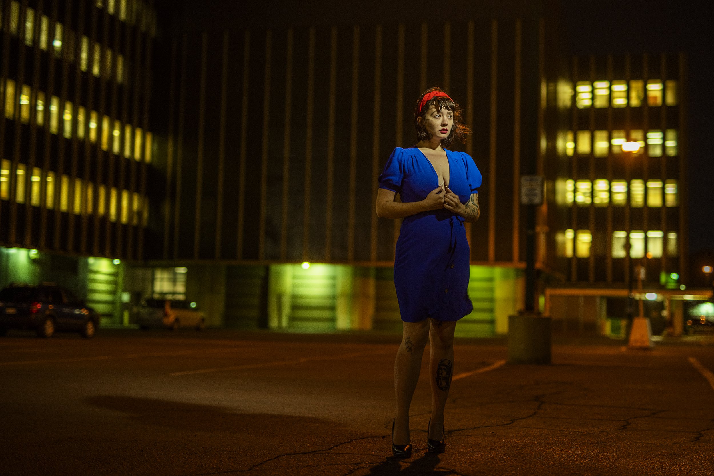 A woman standing in a parking lot at night, wearing a blue dress with a deep neckline, a red headband, and high heels. She has tattoos on her legs and arms and is holding her hands near her chest. The background shows a modern building with lit windo