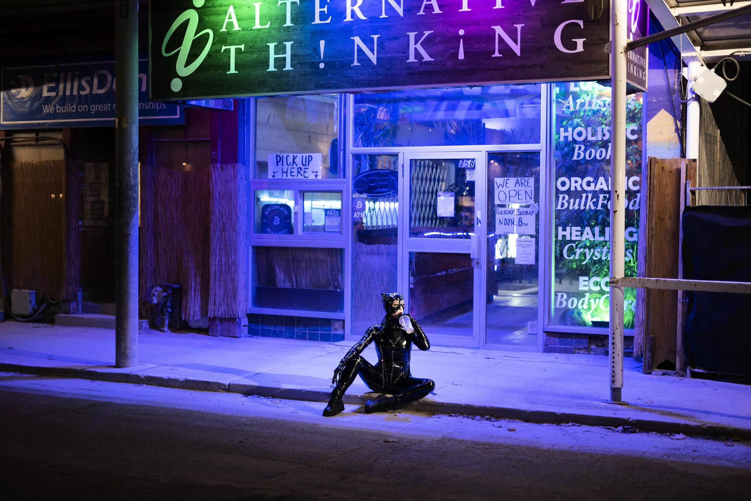 A person dressed as Catwoman in a black, shiny, latex costume sitting on the sidewalk outside a store, drinking from a cup at night with neon signs and store windows illuminated behind.