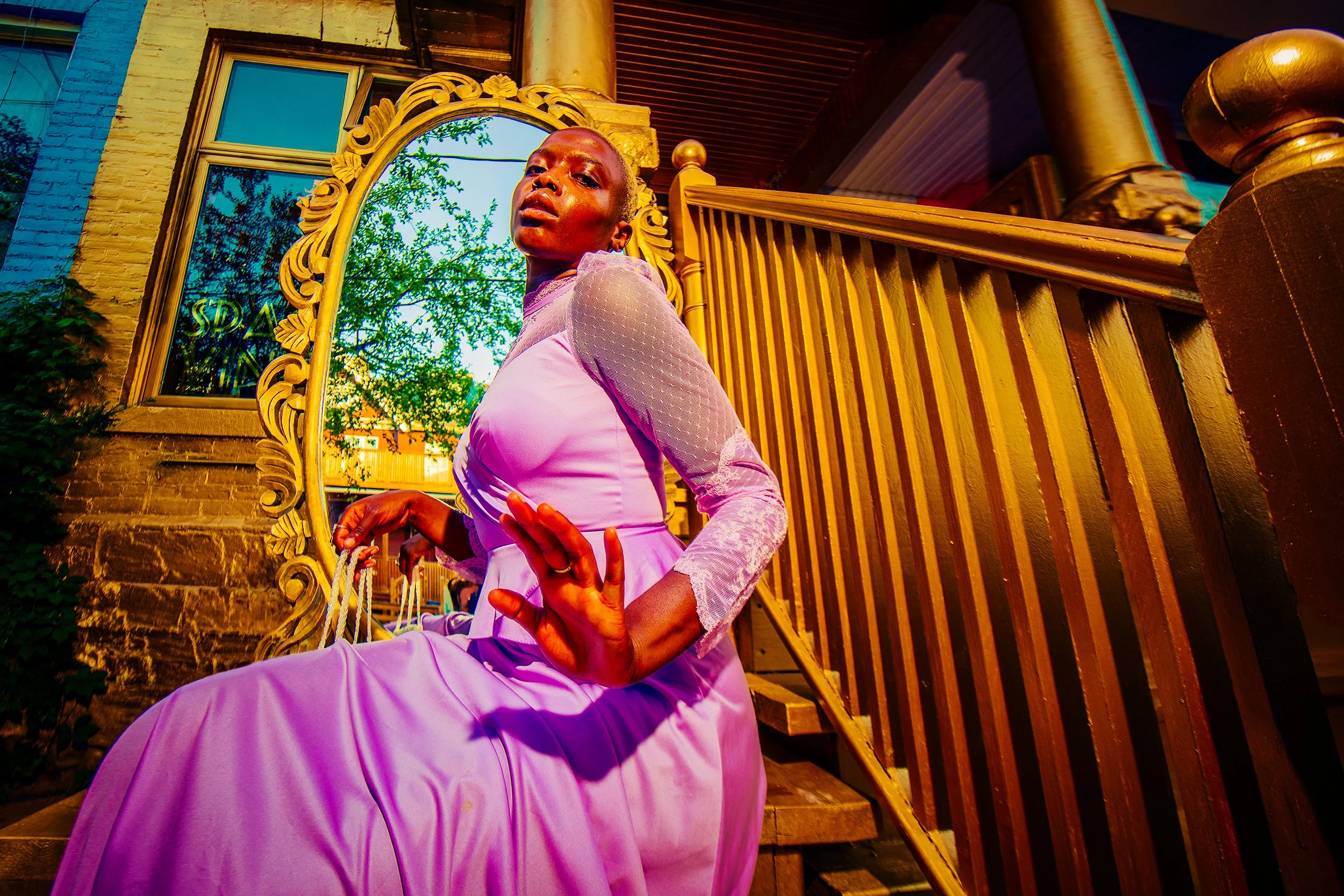 A person dressed in a pink dress with lace details, standing outdoors near a wooden fence and gold-framed mirror, with a yellow brick building and green trees reflected in the mirror.