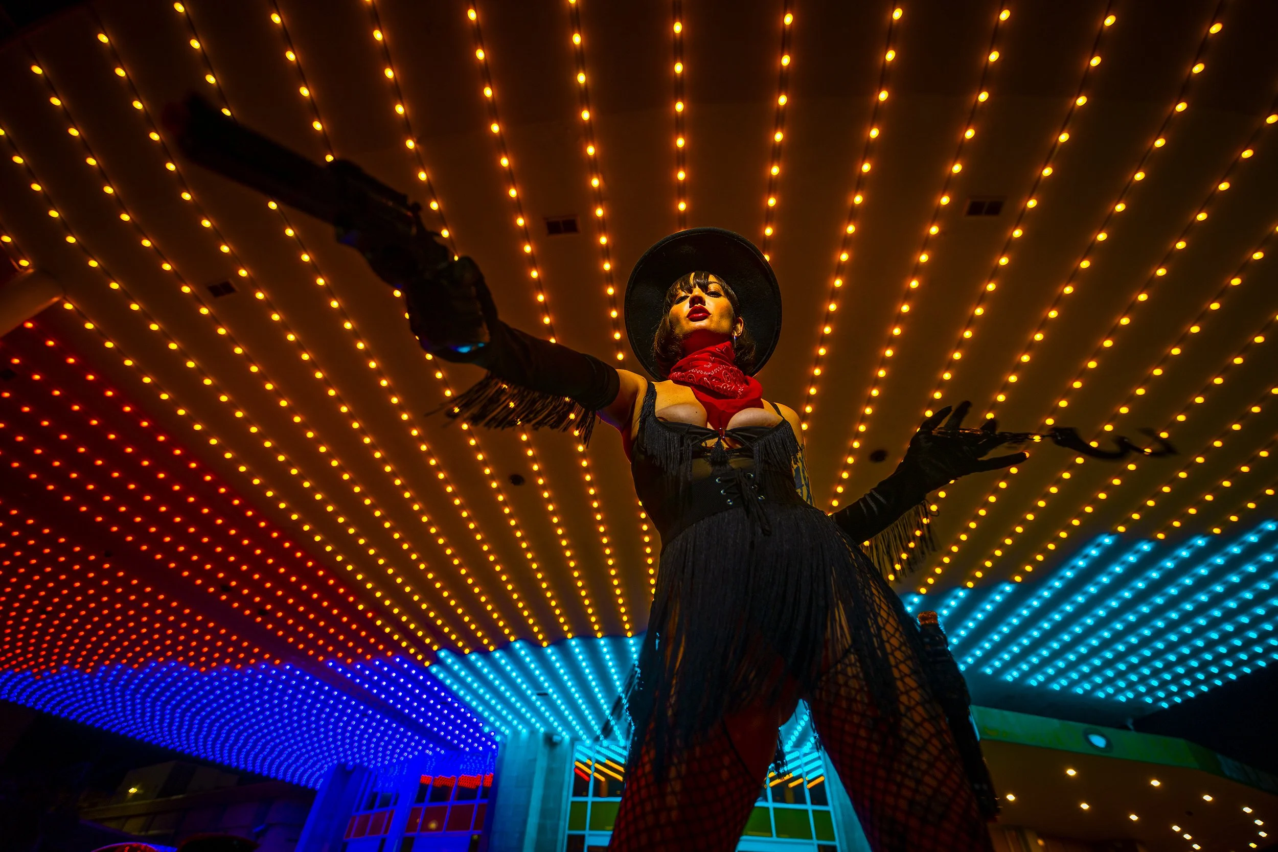 A woman dressed in a black fringed outfit, wearing gloves, a wide-brimmed hat, and a red bandana around her neck, standing beneath a ceiling of vibrant orange and blue LED lights, striking a pose with arms outstretched at night.