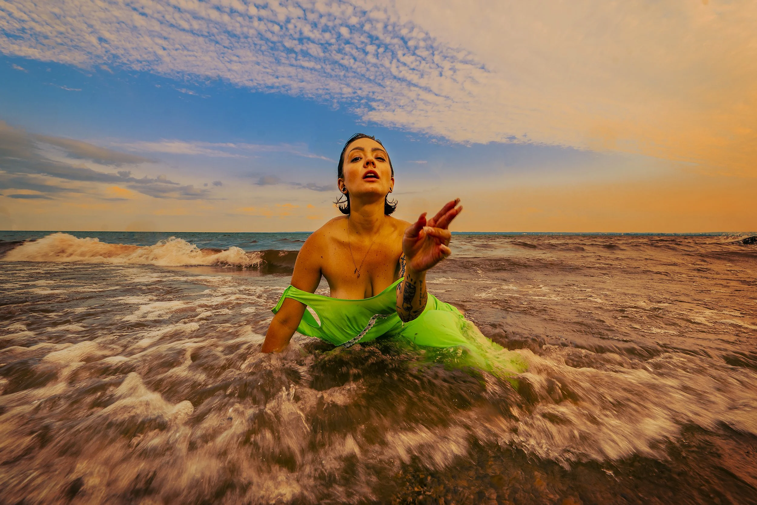 A woman with tattoos on her arm in a green dress is sitting in the ocean at sunset, making a gesture with her hand.