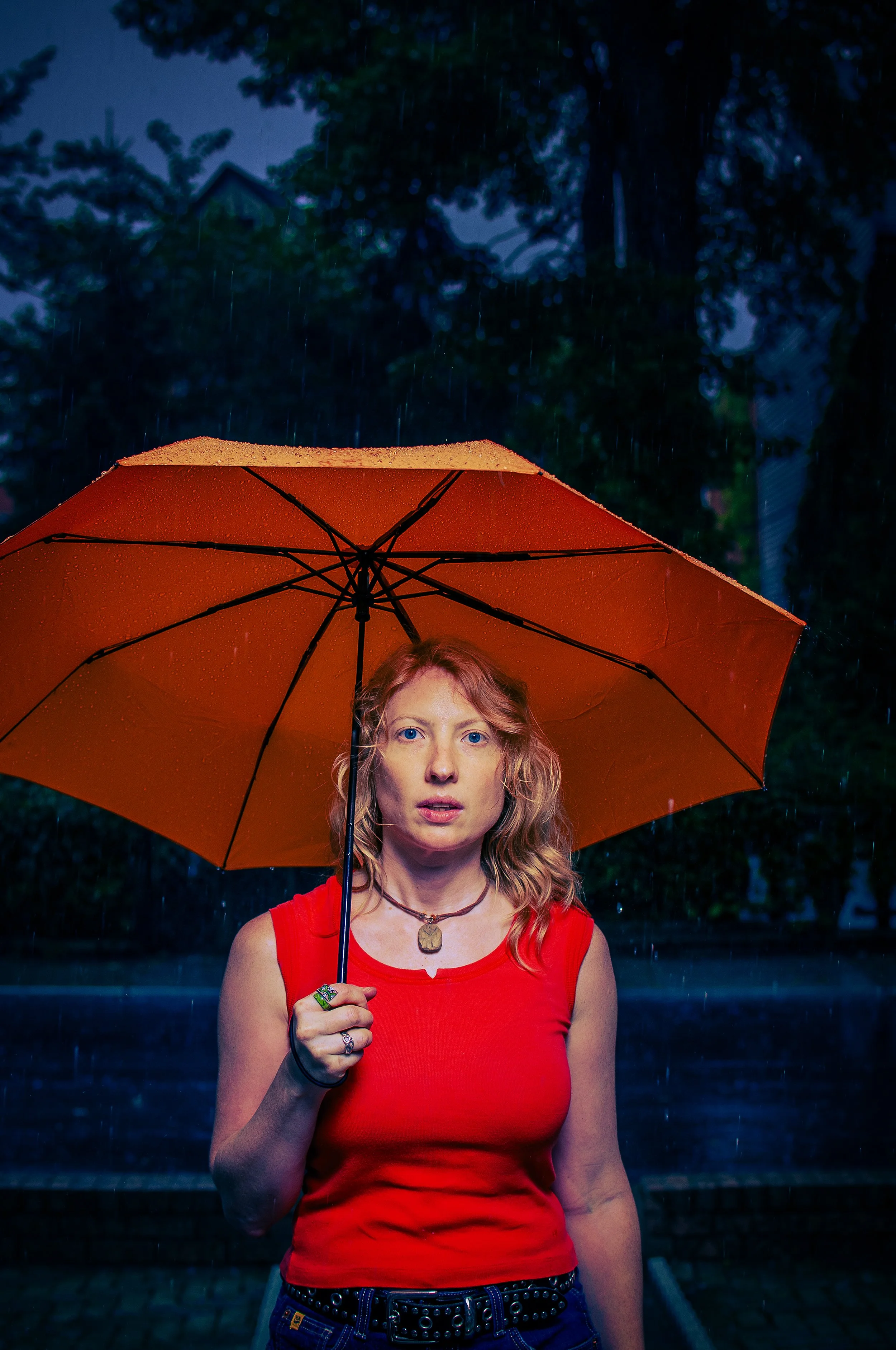 A woman with curly red hair, blue eyes, wearing a red sleeveless top, holding an orange umbrella in the rain during evening or night, with trees and a dark sky in the background.