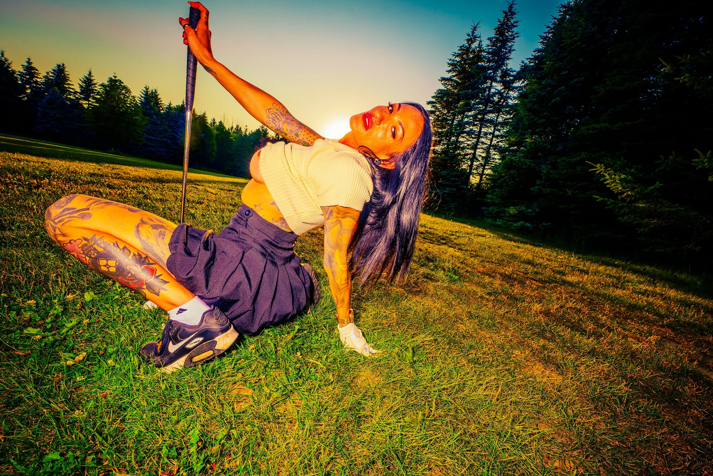 A woman with tattoos on her arms and legs, wearing a yellow crop top, black shorts, and sneakers, posing on the grass with one leg bent and holding a golf club at sunset in a forested area.