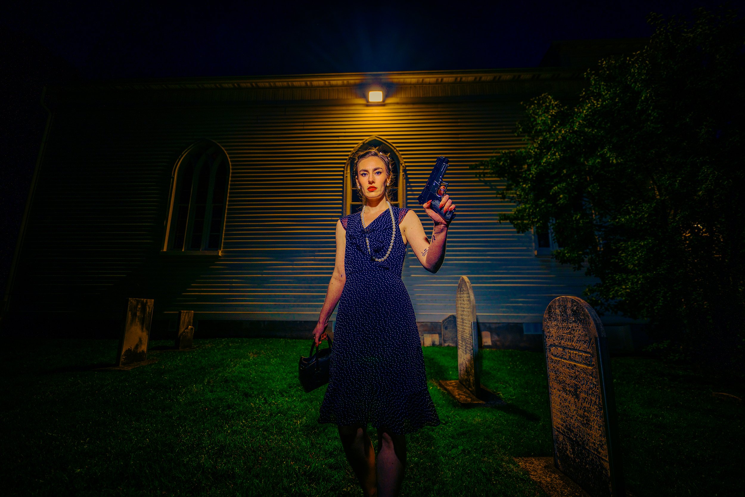 A woman in a vintage polka dot dress with jewelry, holding a gun and a handbag standing outdoors at night, illuminated by a wall light, with old tombstones nearby and a house in the background.
