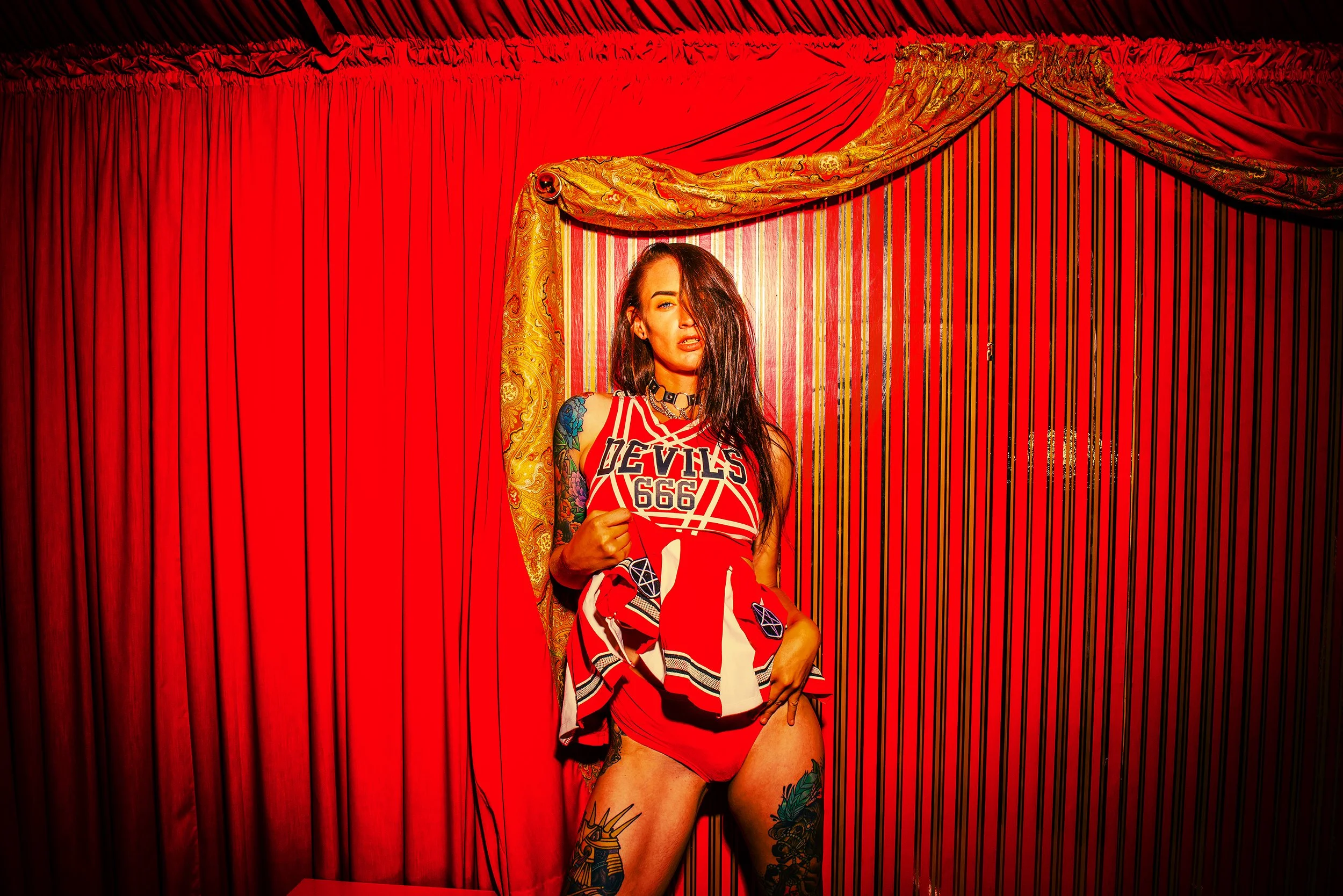 A woman standing in front of red curtains, wearing a sports jersey that says 'Devils 666' and red shorts. She has tattoos on her arms and legs, and long dark hair covering part of her face.