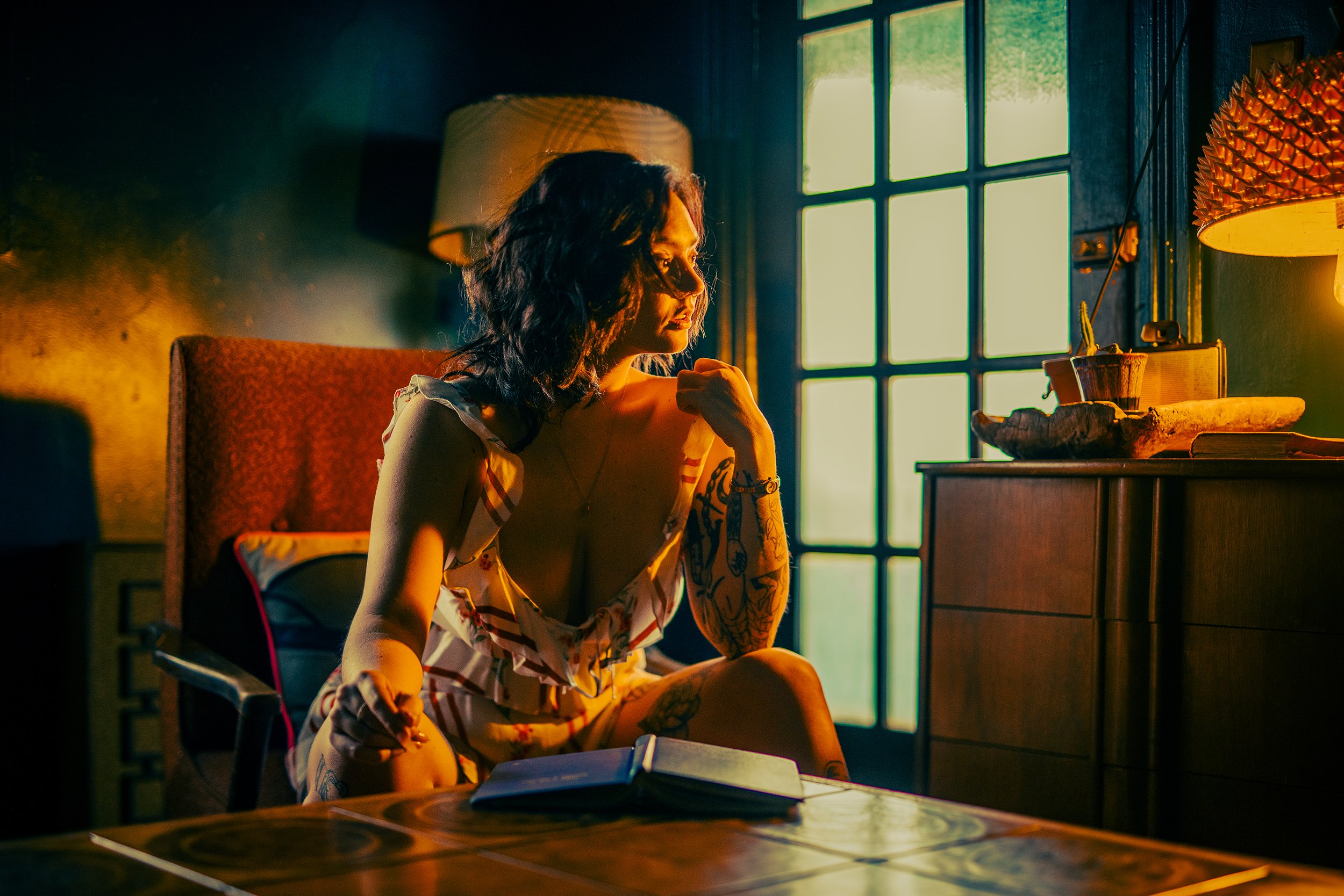 A woman with dark wavy hair and tattoos sitting at a wooden table, glowing from warm natural light coming through large window, in a cozy dimly lit room.