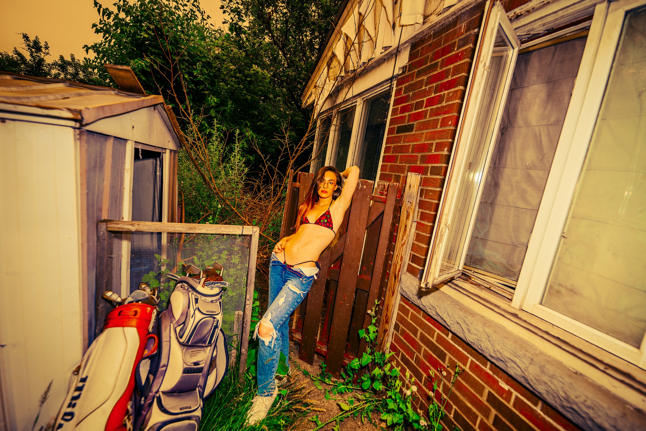 A young woman with long, dark hair, wearing glasses, a black tank top, and ripped jeans, standing outdoors at dusk or nighttime near a brick house with open windows. She is posing with one arm behind her head and the other resting on her waist, surro