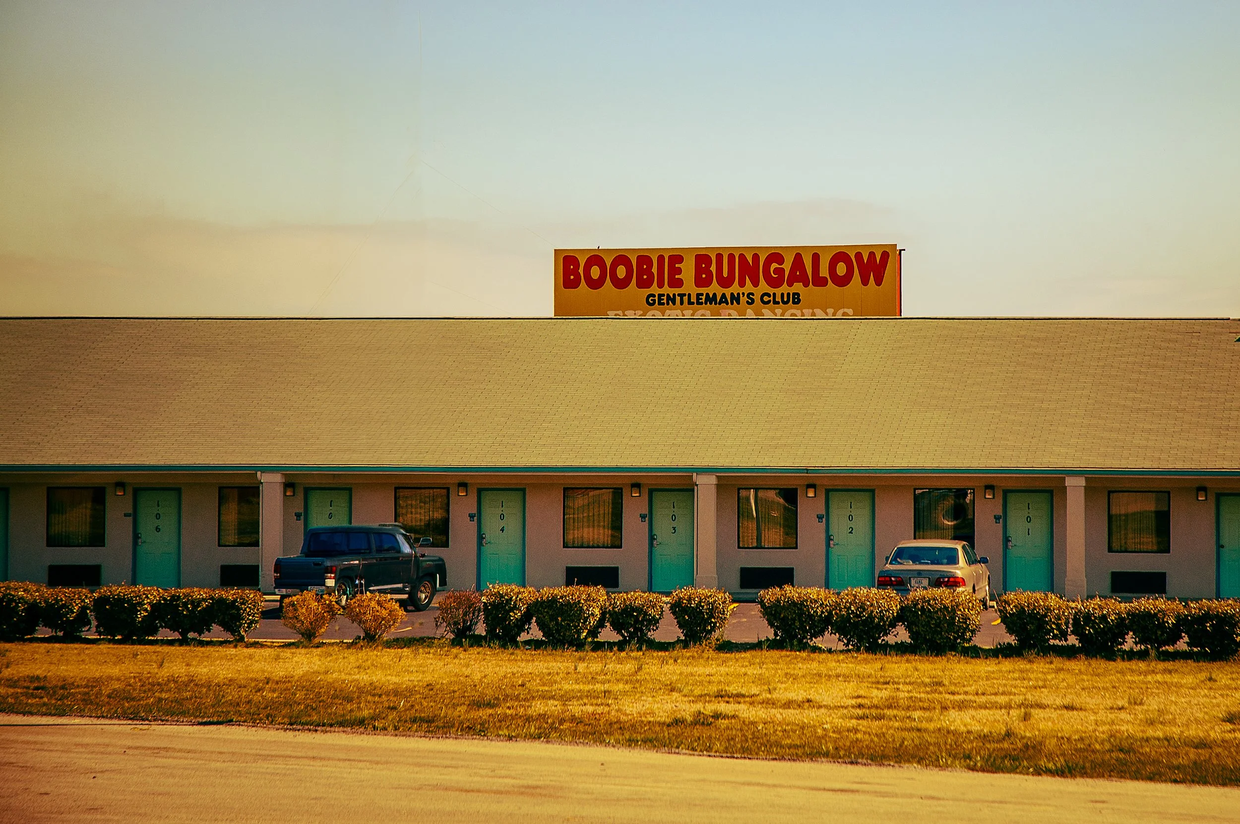 A building with a sign that reads 'Boobie Bungalow' and 'Gentleman's Club' underneath. The building has a yellow roof, green doors, and several cars parked in front. There are bushes and grass in the foreground.