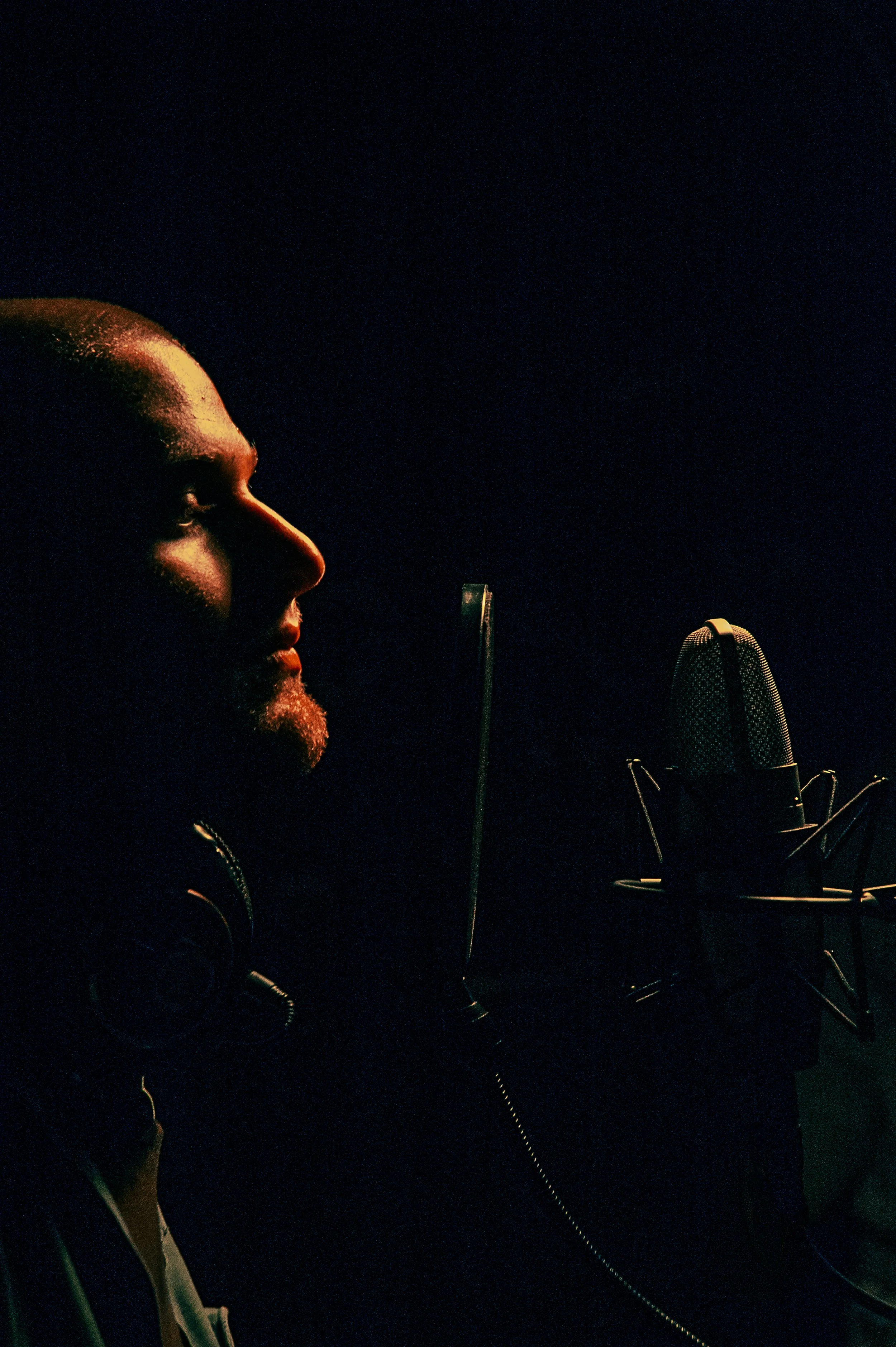 A man with a beard and closed eyes singing into a microphone in a dark recording studio.