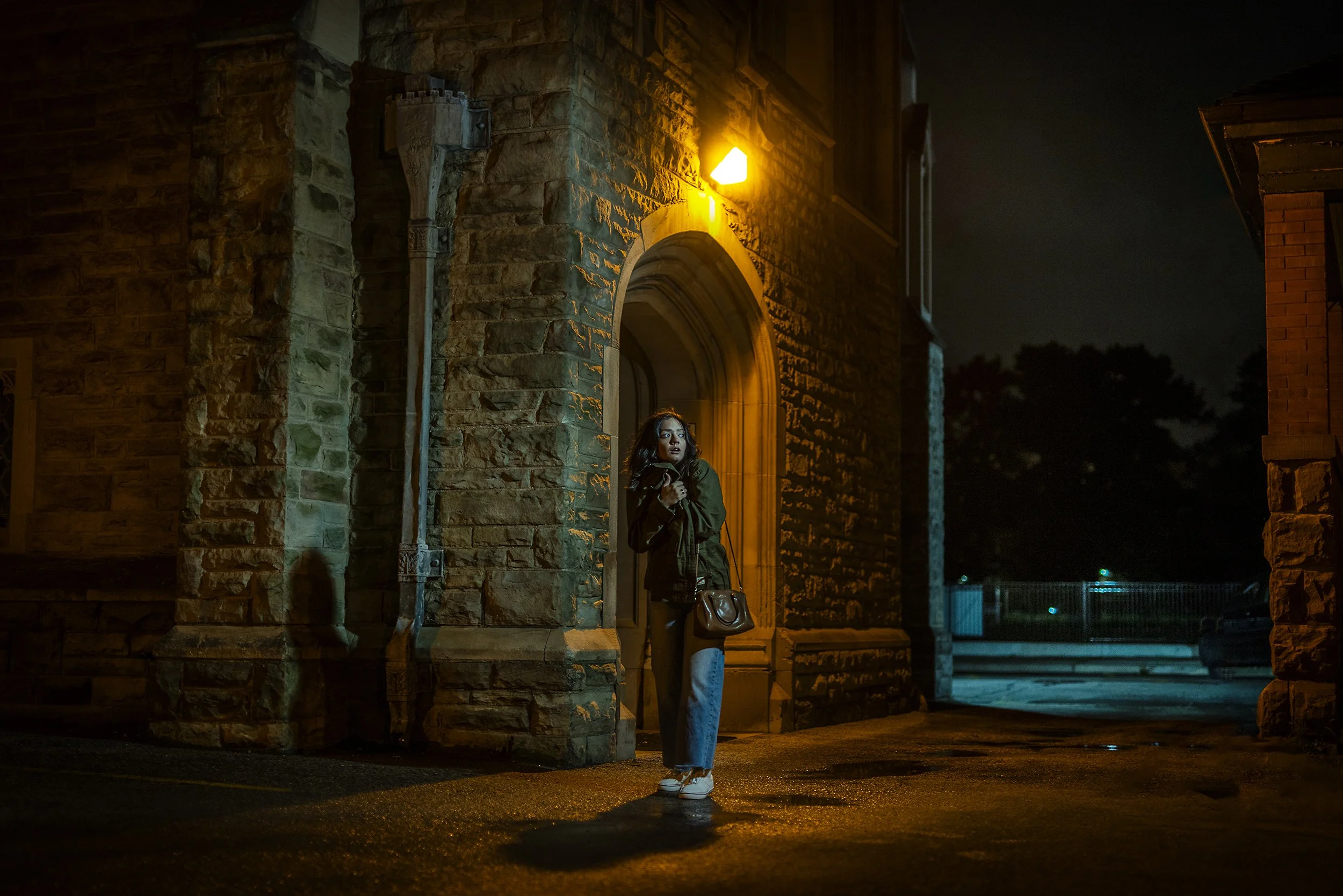 Woman standing under a warm yellow street light outside a stone building at night, holding a small purse, with a dark sky and trees in the background.